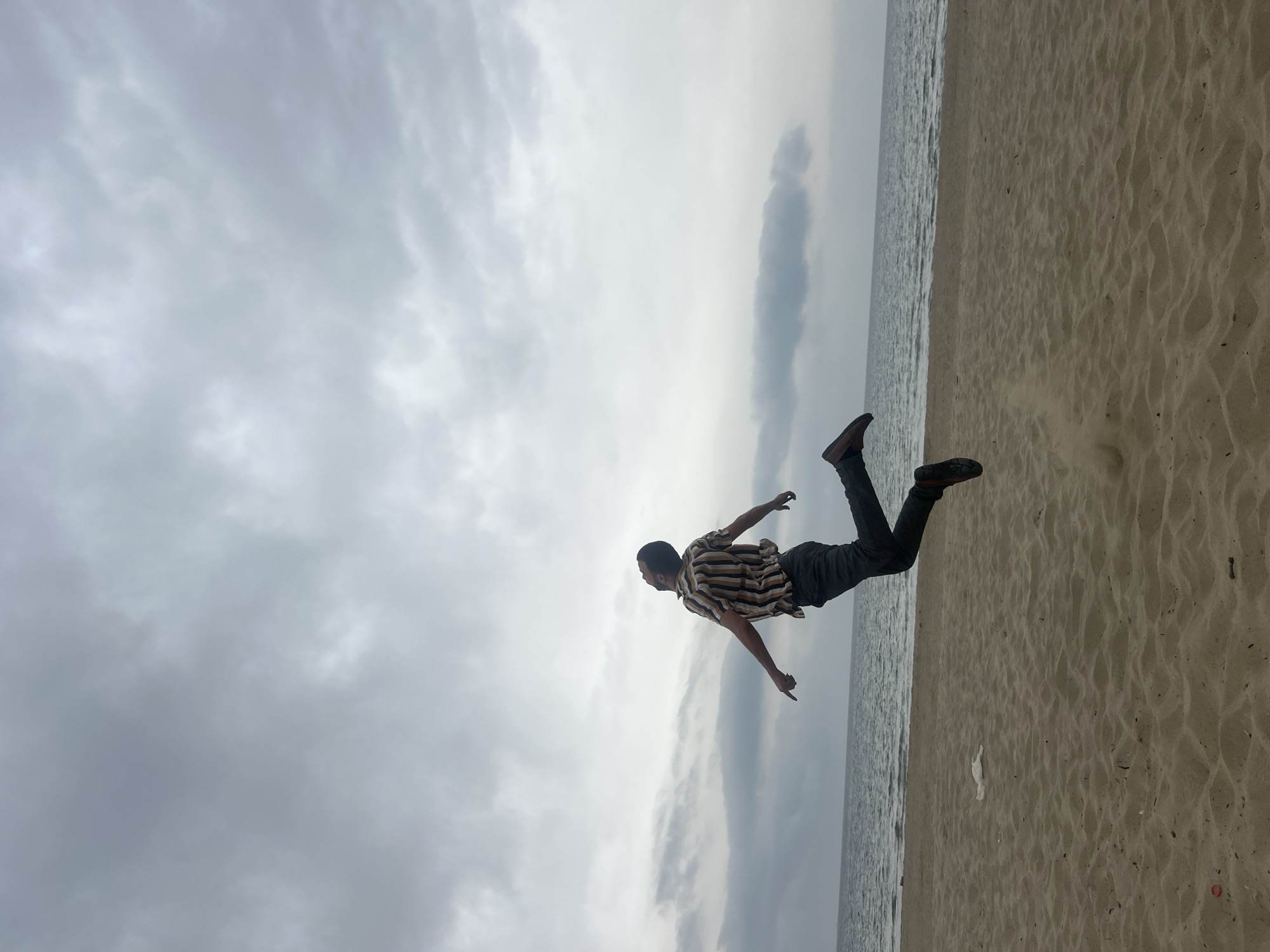 A wide shot captures a beach scene on an overcast day, featuring a man in mid-air against an expansive sky and sea. The image, when oriented correctly, shows the sandy beach in the lower portion of the frame, the sea stretching horizontally across the middle, and the vast, cloudy sky filling the upper half.
The main subject is a man, positioned horizontally across the mid-left to mid-right of the frame. He appears to be jumping or floating, with his body parallel to the horizon line. His arms are outstretched to his sides, and his legs are bent. He is wearing a short-sleeved, striped shirt (tan, brown, and off-white stripes) and dark trousers with dark shoes. His posture conveys a sense of freedom, playfulness, or weightlessness.
The setting is a beach, likely in Martil, Morocco. The foreground and midground consist of light brown sand, showing natural ripples and textures, extending to meet the calm, grey-blue water of the sea. The sea itself is largely placid, reflecting the muted light. Above the sea, a broad, overcast sky dominates the background, filled with soft, grey and white clouds. The heavy cloud cover suggests an overcast day with subdued light, as there are no strong shadows. There is no visible text in the image.