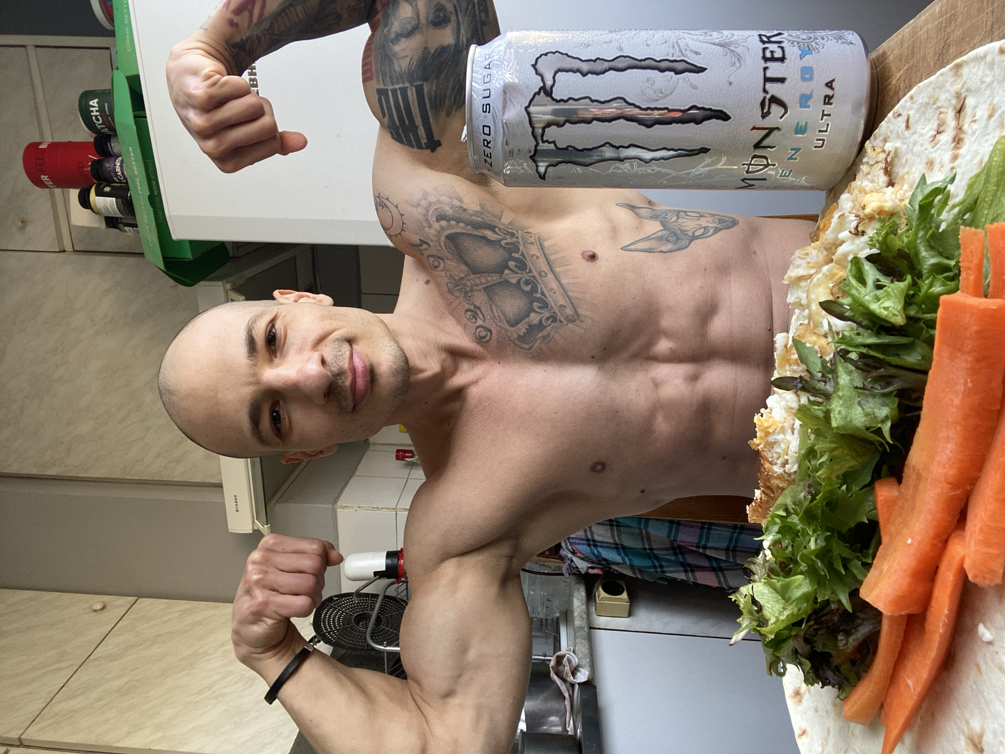 The image depicts an adult male, bald and shirtless, with a muscular physique and visible abdominal muscles, posing in a domestic kitchen. He is flexing both biceps, with clenched fists, and looking directly at the camera with a faint smile. Distinctive tattoos include a large crown design on his left chest and an animal head, possibly a Sphinx cat, on his right lower torso. A black wristband is present on his left wrist.
In the foreground, a meal is arranged on a light-colored wooden surface, likely a cutting board. The meal consists of a large flatbread or tortilla topped with a portion of scrambled eggs or a similar egg preparation, fresh green lettuce leaves, and several thick slices of raw carrots. Adjacent to the meal, a silver can of "Monster Energy Ultra Zero Sugar" is standing upright with its tab opened.
The background reveals a kitchen environment with light-colored, wood-grained upper cabinets and a white tiled backsplash. A "FABER" brand range hood with a glass visor is visible above the man's left shoulder. A white "LIEBHERR" refrigerator is partially visible behind his right arm. On top of the refrigerator, various supplement containers are stacked, including those labeled "MATCHA" and "CARNITINE," along with green packaging bearing text like "DRINK BETTER FEEL BETTER." On the counter to the left, a black circular metal object, a white pump dispenser, and a plaid-patterned cloth are present, with a beige electrical outlet visible on the wall.
The specific location within the city of Pleven, Bulgaria, cannot be determined from the visual information presented in the image.