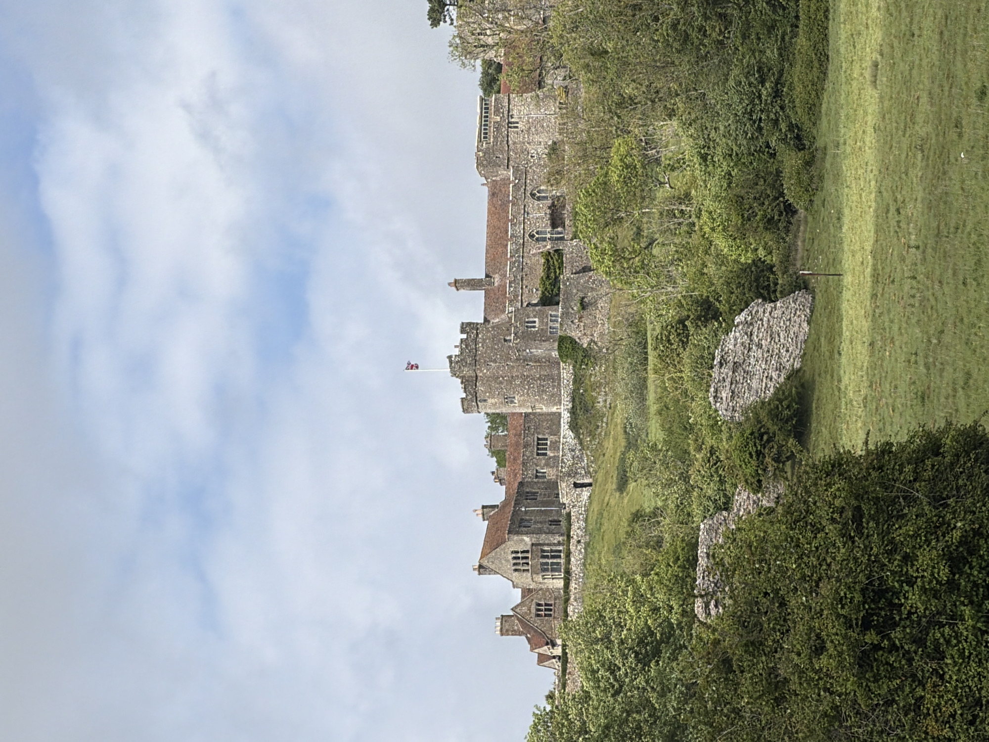 The image depicts Saltwood Castle, situated near Hythe in the Folkestone and Hythe District of Kent, United Kingdom. The castle, constructed of grey stone, features battlements, towers, and residential buildings with red tile roofs. The British flag flies atop one of the towers. The castle is positioned on a hillside, surrounded by lush green vegetation including trees and grass. A field with a partially visible stone structure borders the castle grounds. The sky above exhibits a pale blue color with scattered white clouds.
