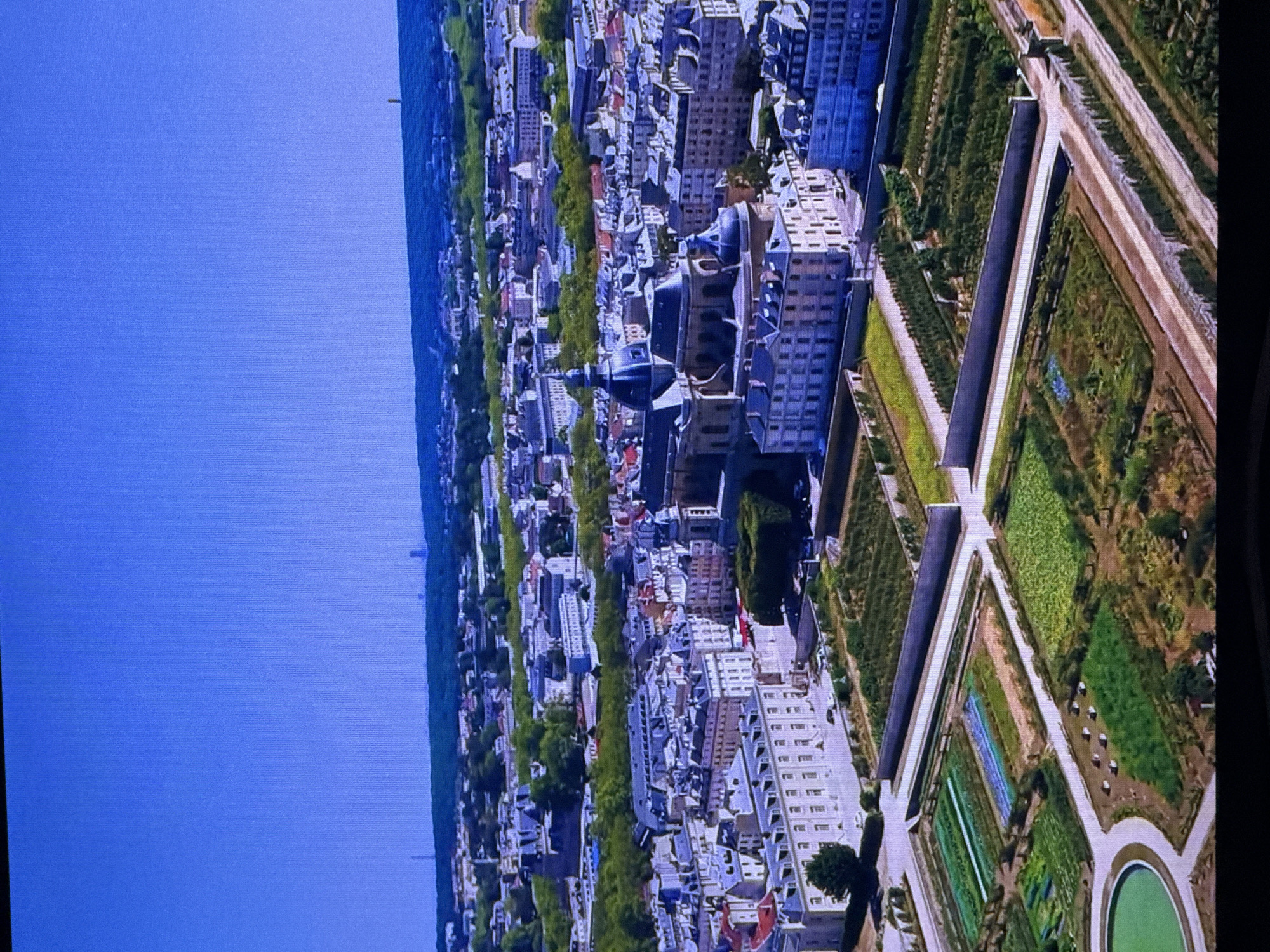 This is an aerial photograph of a city with a large domed building and meticulously maintained gardens in the foreground. The city appears to be European in architecture, with mansard roofs and stone facades. The gardens are divided into geometric plots, with some appearing to be agricultural or horticultural. There is a round pool or fountain on the left side of the garden. The sky is a clear blue. The image does not depict Dubai, United Arab Emirates, nor does it contain any people.