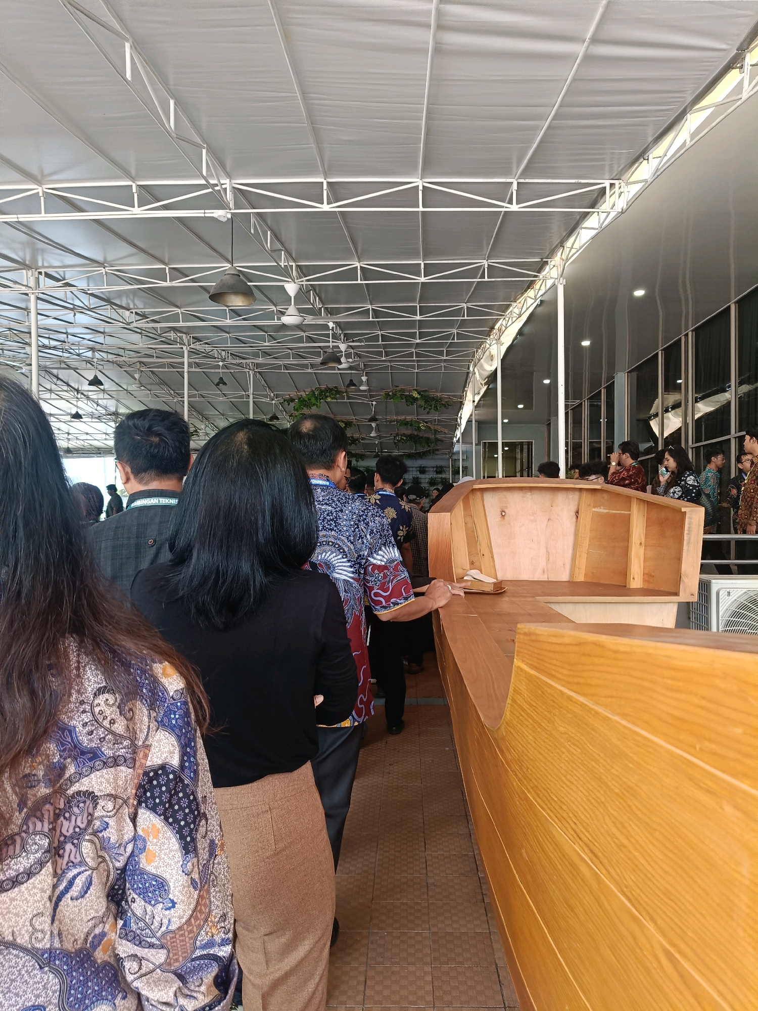 The image shows an indoor event, possibly a ceremony or exhibition, at a venue with a large, open-plan structure with a white, tent-like roof supported by a metal frame. The ceiling also features industrial lighting fixtures and ceiling fans. The event appears to be taking place in North Jakarta, Indonesia, given the batik attire worn by many attendees and the architectural style.
In the foreground, the back of a woman with long, dark hair is visible, wearing a colorful batik patterned top. To her right, another woman in a black top and tan trousers stands. Further down the line, several men and women are seen, many in batik shirts. The group appears to be queuing or observing something.
To the right of the line of people, a prominent wooden structure is visible. It has a curved shape and is constructed from light-colored wood, resembling part of a boat or a display platform. The construction is detailed with visible planks and joints.
In the background, through large glass windows, other attendees can be seen, as well as parts of a building interior. An air conditioning unit is also visible on the right side of the image. The overall impression is of a public gathering in a semi-open or covered outdoor space, possibly a community hall or event center.