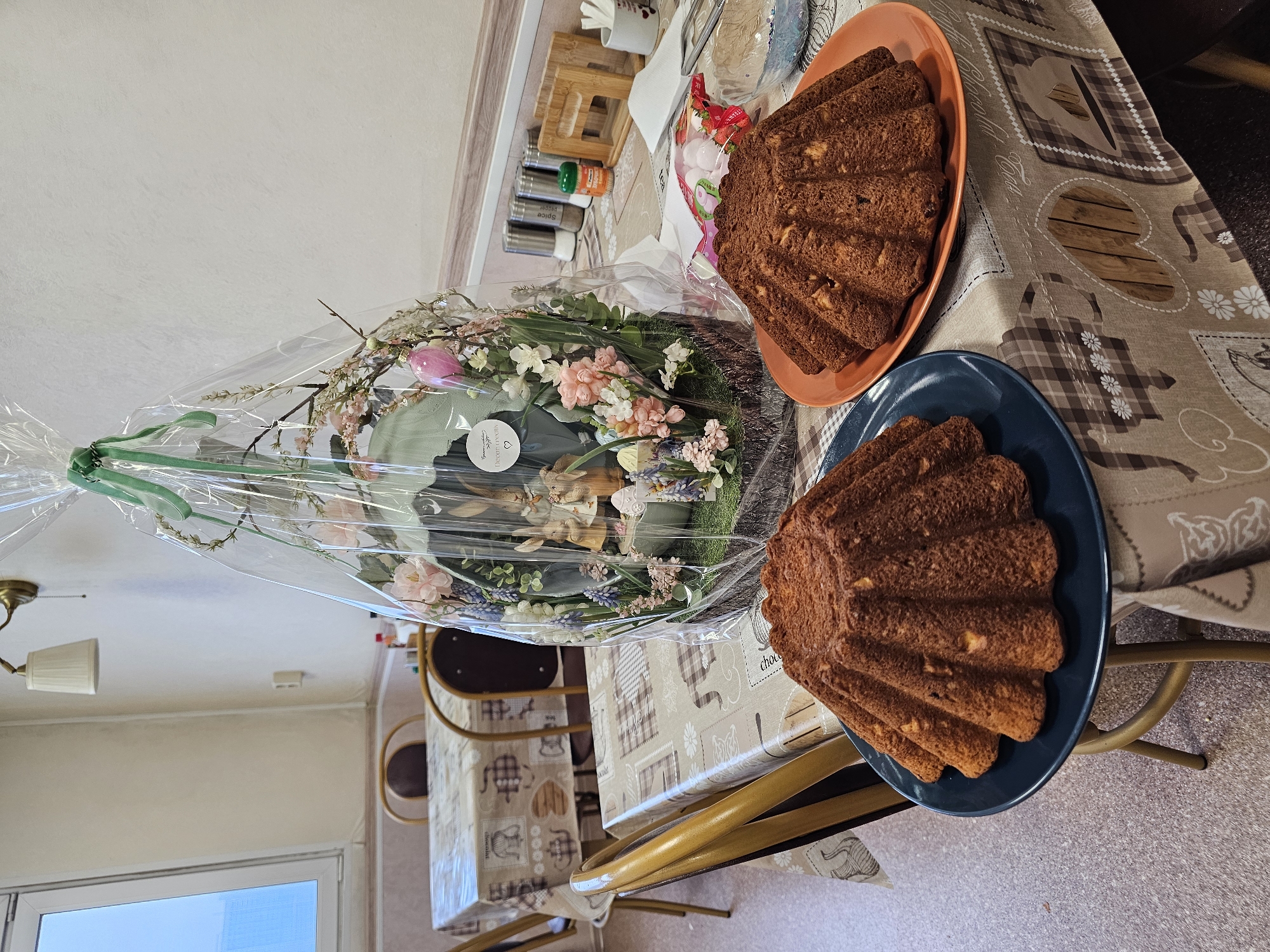 The image captures an indoor scene on a table covered with a patterned tablecloth. Two bundt cakes are displayed on separate plates - one on an orange plate and the other on a blue plate. Next to the cakes is a cellophane-wrapped gift basket containing flowers, easter eggs and other decorative elements. Condiment containers are visible in the background. The photo was likely taken indoors, possibly in a dining area. The location, Khimki, Russia is not discernible from the given information.