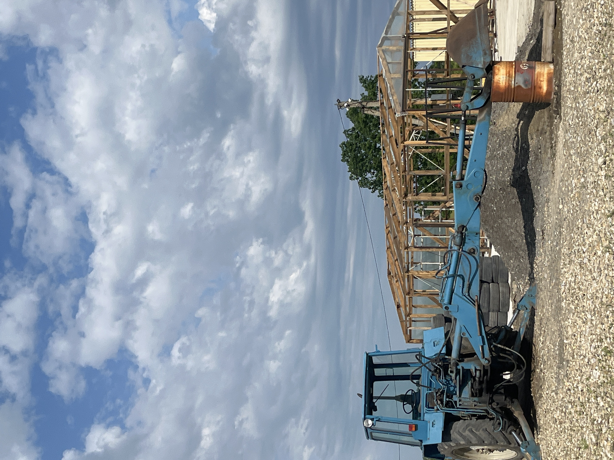 The image shows a blue tractor with a front loader parked on a gravel surface. The tractor is angled towards the right side of the frame. Behind the tractor is a wooden frame structure, possibly under construction or renovation. A rusty metal barrel is positioned near the front loader. The background consists of a cloudy sky. The location, Chetuk, Russia, is provided as context but cannot be verified from the visual information alone. There are no people visible in the image.