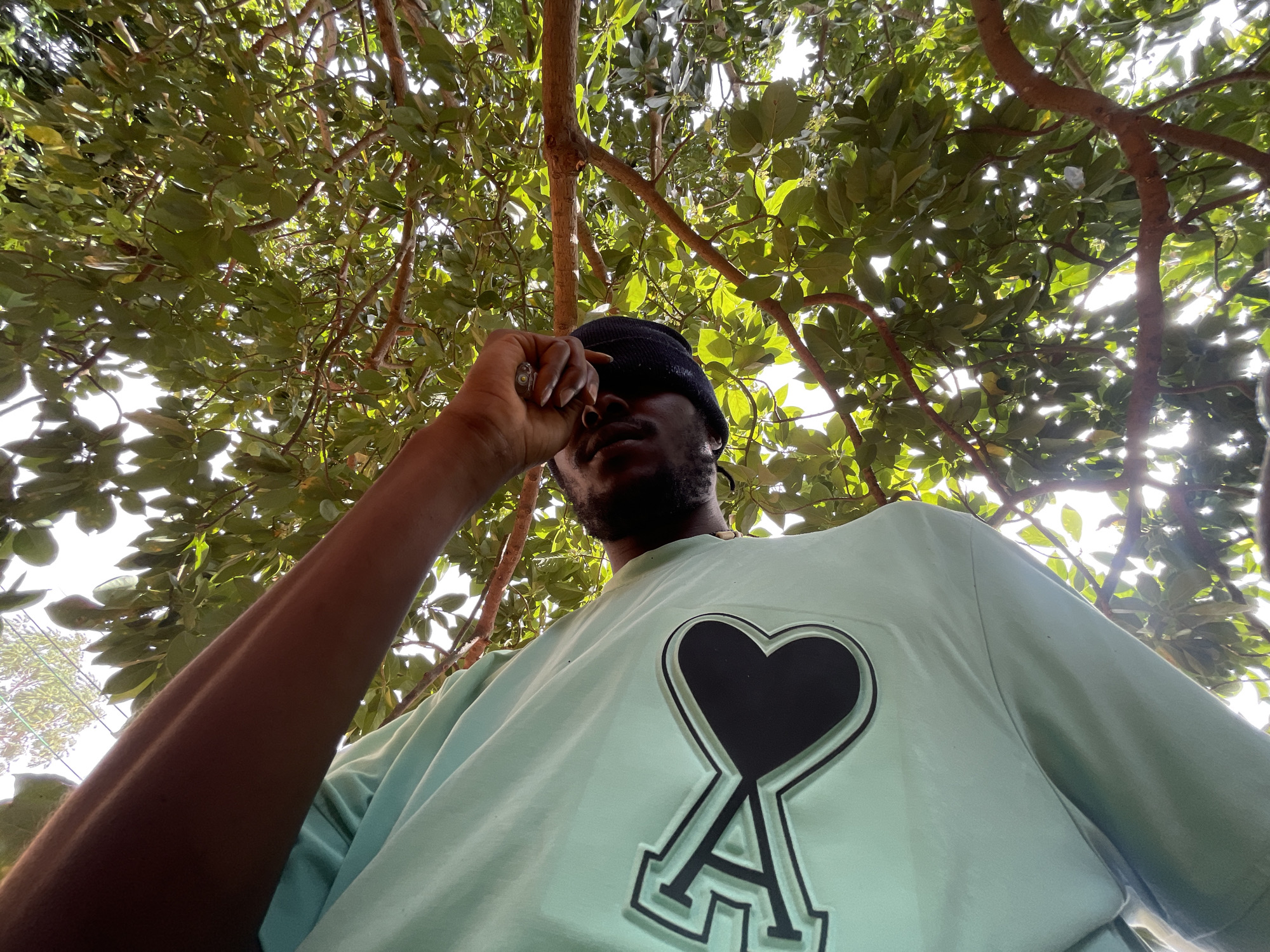 A low-angle shot shows a man under a tree. He is wearing a black beanie and a light blue t-shirt with a black heart and a stylized letter "A" design on the chest. His arm is raised, hand near his forehead. The background consists of tree branches and leaves against a bright sky. The scene is likely located in Jos, Nigeria.