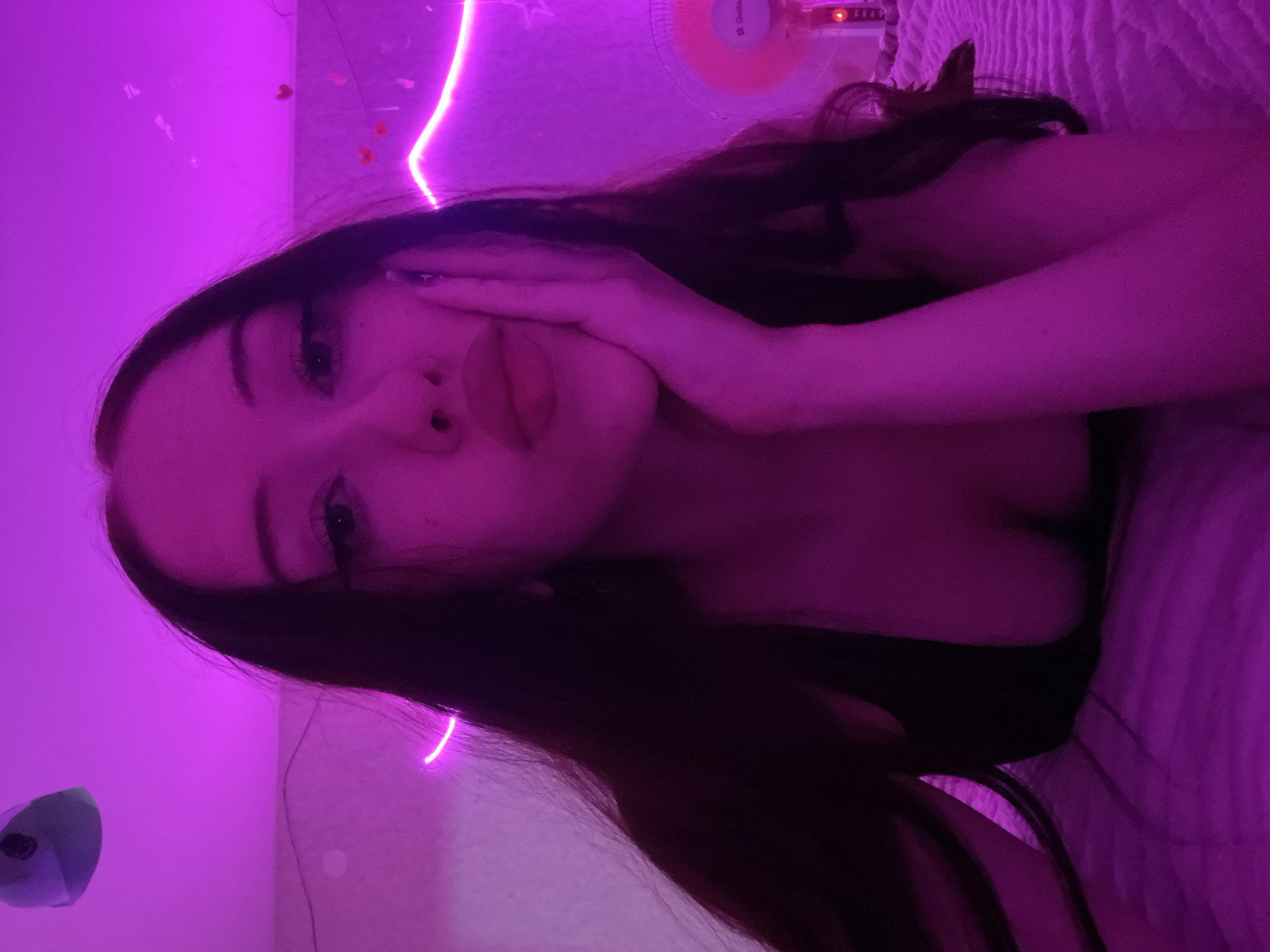A young woman with long, dark hair is lying on a bed, illuminated by pink neon lights. She is looking directly at the camera, with her hand resting on her cheek. Her lips are slightly parted. She is wearing a dark top that reveals her cleavage. In the background, there are more pink neon lights, some heart-shaped decorations, and a pink fan. The room appears to be a bedroom. There is no information in the image to suggest the location is Votkinsk, Russia.
