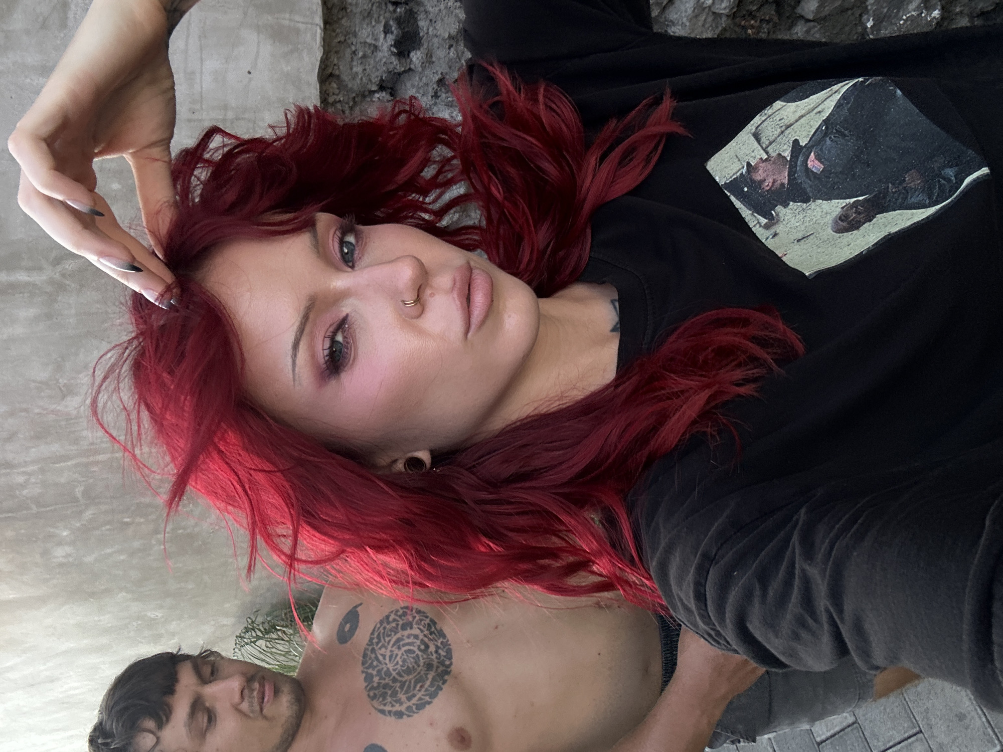 Two individuals are posing for a photograph outdoors. The person in the foreground is a woman with vibrant red hair, wearing makeup and a black t-shirt with a graphic print. She is touching her hair with her right hand. To her left and slightly behind her is a man with short dark hair, shirtless, displaying tattoos on his chest. The background consists of a textured wall and a stone surface, suggesting an urban or rustic outdoor setting. There is no discernible indication of the specific location within North Kuta, Indonesia.