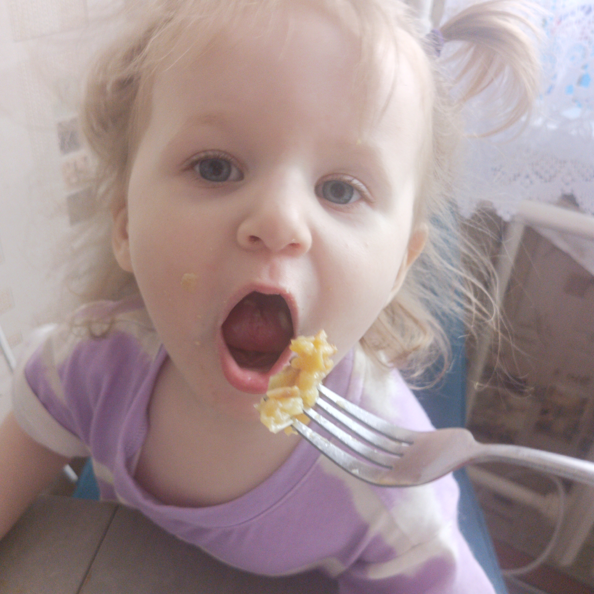 A young, light-skinned child with blond hair in pigtails is in the act of eating. The child is wearing a purple and white striped shirt. A fork with some food on it is entering the child's open mouth. There appears to be some food on the child's face. The background is blurred, but suggests an interior space with kitchen elements. It is not possible to confirm the location is Chaikovsky, Russia from this image.