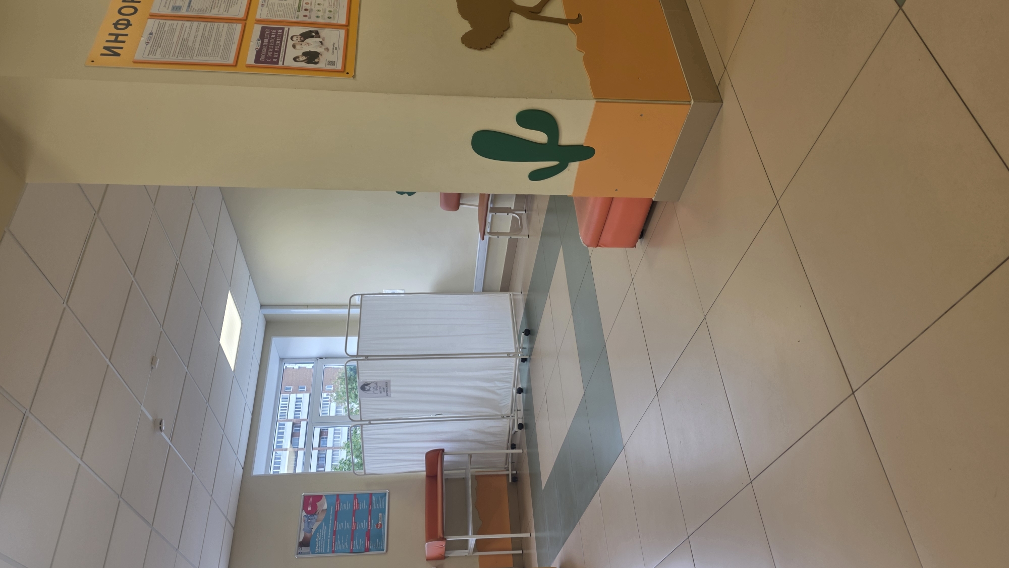 A photograph taken indoors at a healthcare facility in Saint Petersburg, Russia. The area features tiled floors and ceiling, suggestive of an institutional setting. The interior space is illuminated, likely by a combination of natural light coming in through a window on the left side of the scene and artificial light from ceiling fixtures.
The room contains several pieces of furniture, including a foldable privacy screen, likely for medical examinations, and a waiting area with benches. A wall is decorated with informational posters and artwork, including what appears to be a desert-themed mural.
The photograph appears to be taken from a stationary position, looking slightly upwards towards the left side of the room. There are no people visible in the photograph.