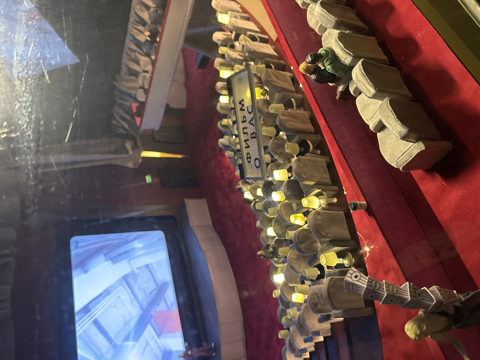 The image shows a miniature diorama of a theater in Moscow, Russia. The diorama depicts theatergoers seated in rows of plush seats, illuminated by small lights. The sign "О ФИЛЬМ ВАС" (O FILM VAS) is visible. A television screen on the left displays abstract white and blue patterns. There are miniature figures of theatergoers in the seats and on the red carpeted floor. The diorama is enclosed in a glass case, visible by the reflections and water droplets on the glass.
