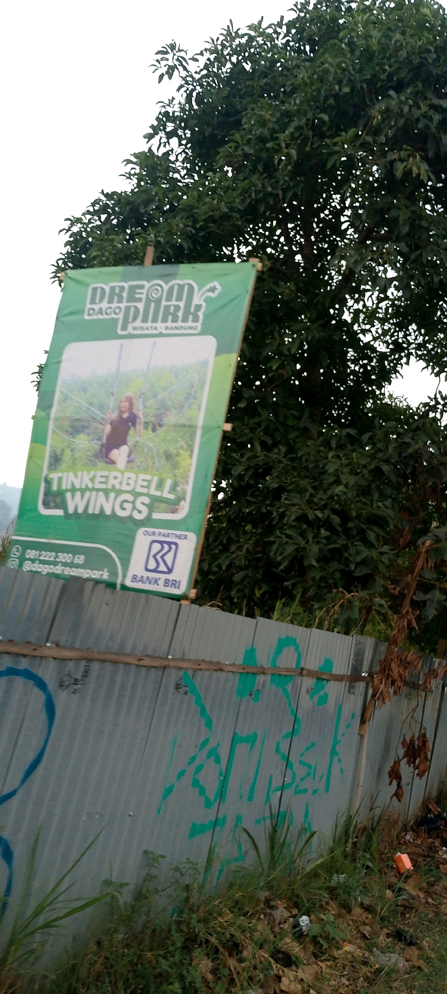 A green banner for "DREAM PARK" is displayed outdoors, angled slightly to the left and supported by wooden poles. The banner features an image of a young woman on a swing, with the text "TINKERBELL WINGS" below her. Towards the bottom left, a phone number "081 222 300 68" and an Instagram handle "@dogodreampark" are visible. To the right of this, a Bank BRI logo and the text "OUR PARTNER" are present. The banner is positioned against a backdrop of lush green trees and an overcast sky. In the foreground, a corrugated metal fence runs horizontally across the image, partially obscured by grass and debris. Graffiti in green paint is visible on the fence, and a small orange object, possibly trash, lies on the ground near the fence. The scene appears to be located in a park or outdoor recreational area, with the presence of trees and vegetation suggesting a natural setting. The overall impression is that of an advertisement for an amusement park. Based on the visible information, the location can be identified as Mekarwangi, Indonesia, likely at Dream Park Dago.