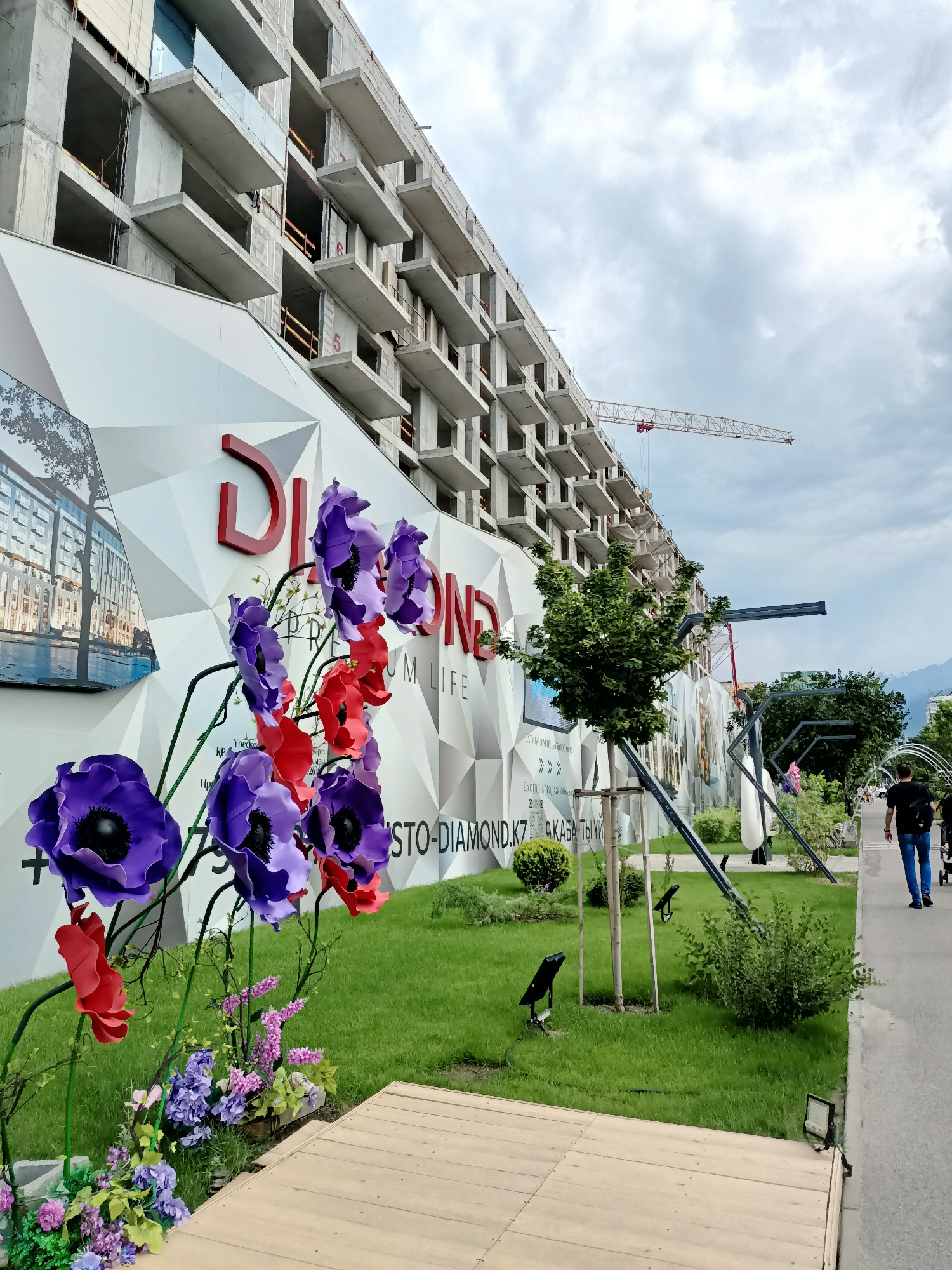 The image depicts a construction site in Almaty, Kazakhstan, characterized by a large, unfinished multi-story building made of concrete and scaffolding. In the foreground, oversized artificial flowers, predominantly purple and red, are displayed. A white, geometrically patterned wall with red lettering that reads "DI AMOND" and text in Cyrillic script is situated in front of the construction. The area also features a paved walkway on the right, with a person walking away from the camera, and a grassy area with trees and decorative lighting. In the background, a construction crane is visible, and a cloudy sky suggests overcast weather. The scene appears to be a promotional or display area adjacent to a construction project, possibly a residential complex.
