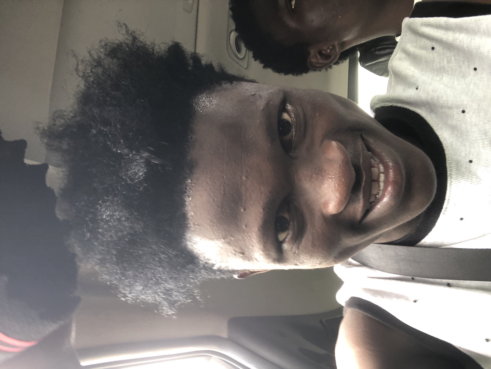 A young Black male is captured in a close-up, selfie-style shot, looking directly at the camera with a wide smile. His hair is dark and stands out in a voluminous, natural afro. He is wearing a white sleeveless shirt with black trim and appears to be inside a vehicle, evidenced by the presence of a seatbelt strap and what seems to be part of a car interior in the background. Another individual's profile is visible to the right of the main subject. The lighting suggests it might be daytime, possibly with some sunlight filtering in. The context of Ado, Nigeria, places this scene within that specific geographical and cultural setting.