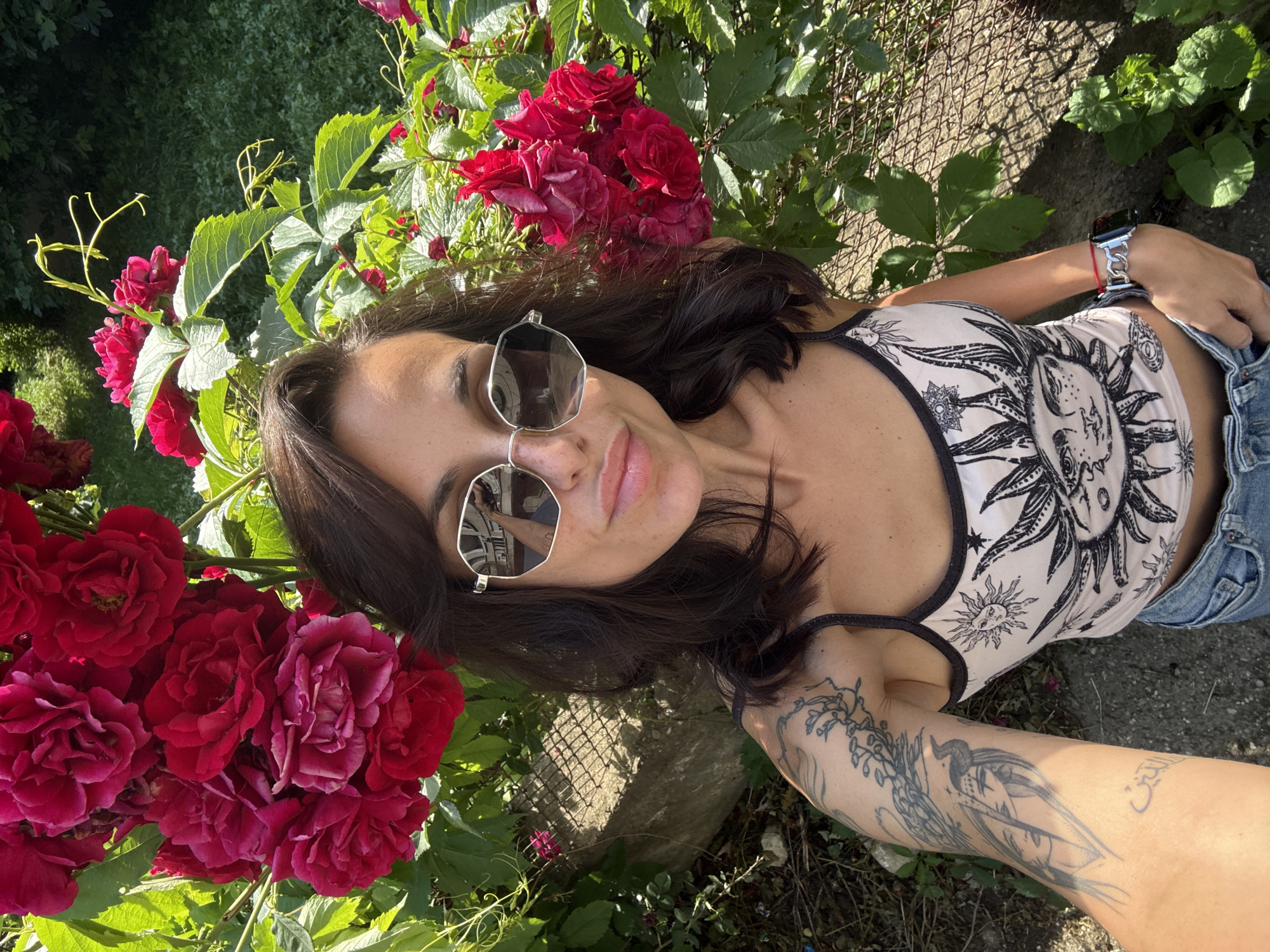 The image is a selfie taken outdoors, likely in a garden. A woman with dark hair and sunglasses is positioned in the center, angled towards the camera. She is wearing a tank top with a celestial design and denim shorts. Her right arm is visible and features tattoos.
A vibrant display of red roses is prominently featured to the left. A chain-link fence and surrounding vegetation are visible in the background. The photo appears to have been taken in Pleven, Bulgaria.