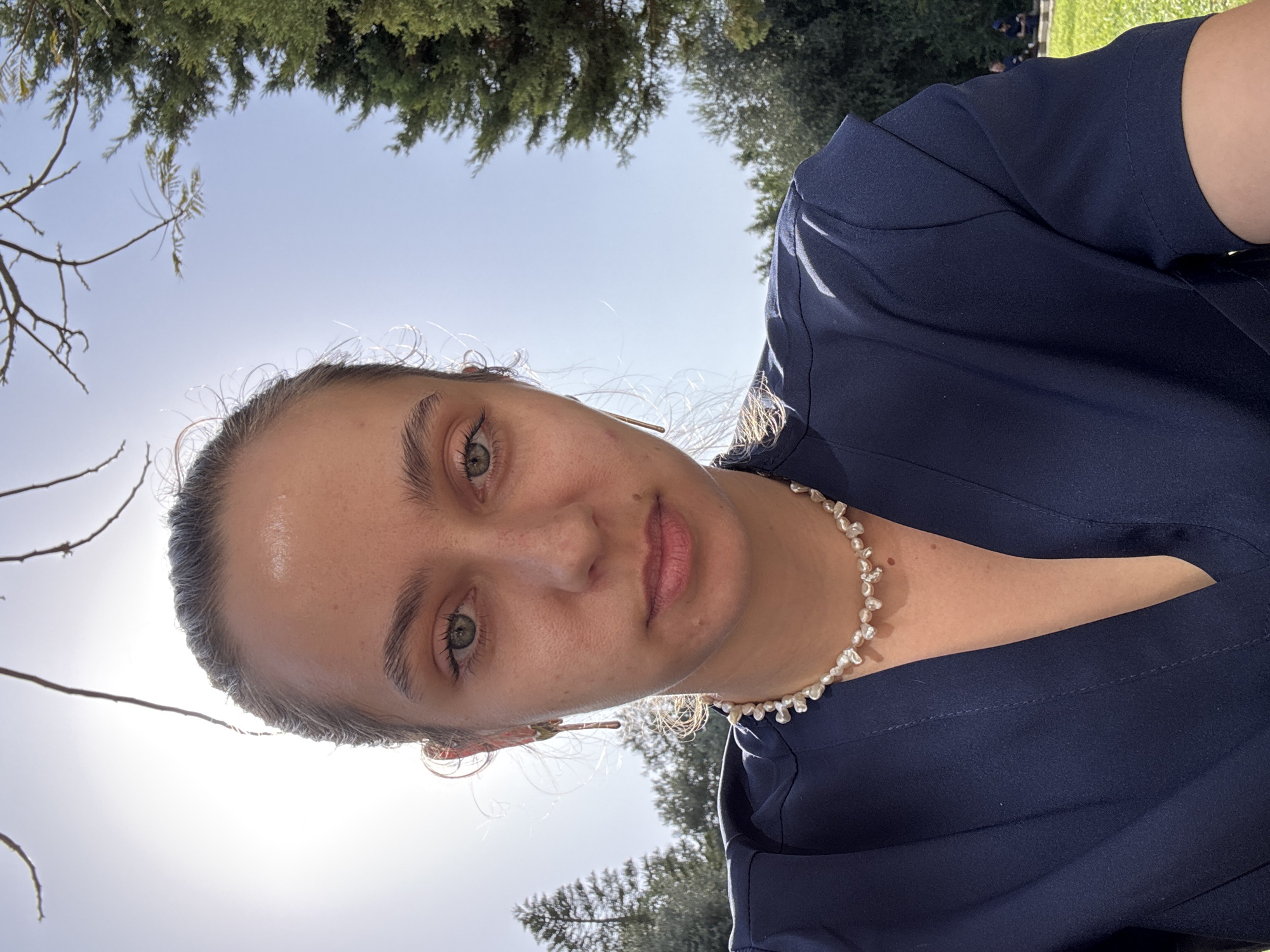 The image, which appears to be a rotated selfie, features a young woman outdoors under bright daylight. When correctly oriented, she is looking directly at the camera with a neutral expression. Her head and upper torso are prominent, wearing a dark blue top, possibly scrubs or a simple blouse. Around her neck is a necklace composed of small, irregular white beads, resembling freshwater pearls. She has light skin, green eyes, and her hair, which is light brown or dark blonde, is mostly pulled back, with some loose, curly strands around her face.
The lighting is bright and sunny, with the sun appearing to be behind and to her left, creating highlights on her hair and shoulder. The background features a clear, light blue sky. On the upper right, lush green foliage from trees is visible, while on the left, bare branches extend into the frame. A patch of green grass and blurred shapes in the far background on the right suggest an outdoor setting like a park or campus, consistent with a location such as Tel-Aviv, Israel. The weather is clear and sunny. There is no visible text in the image.