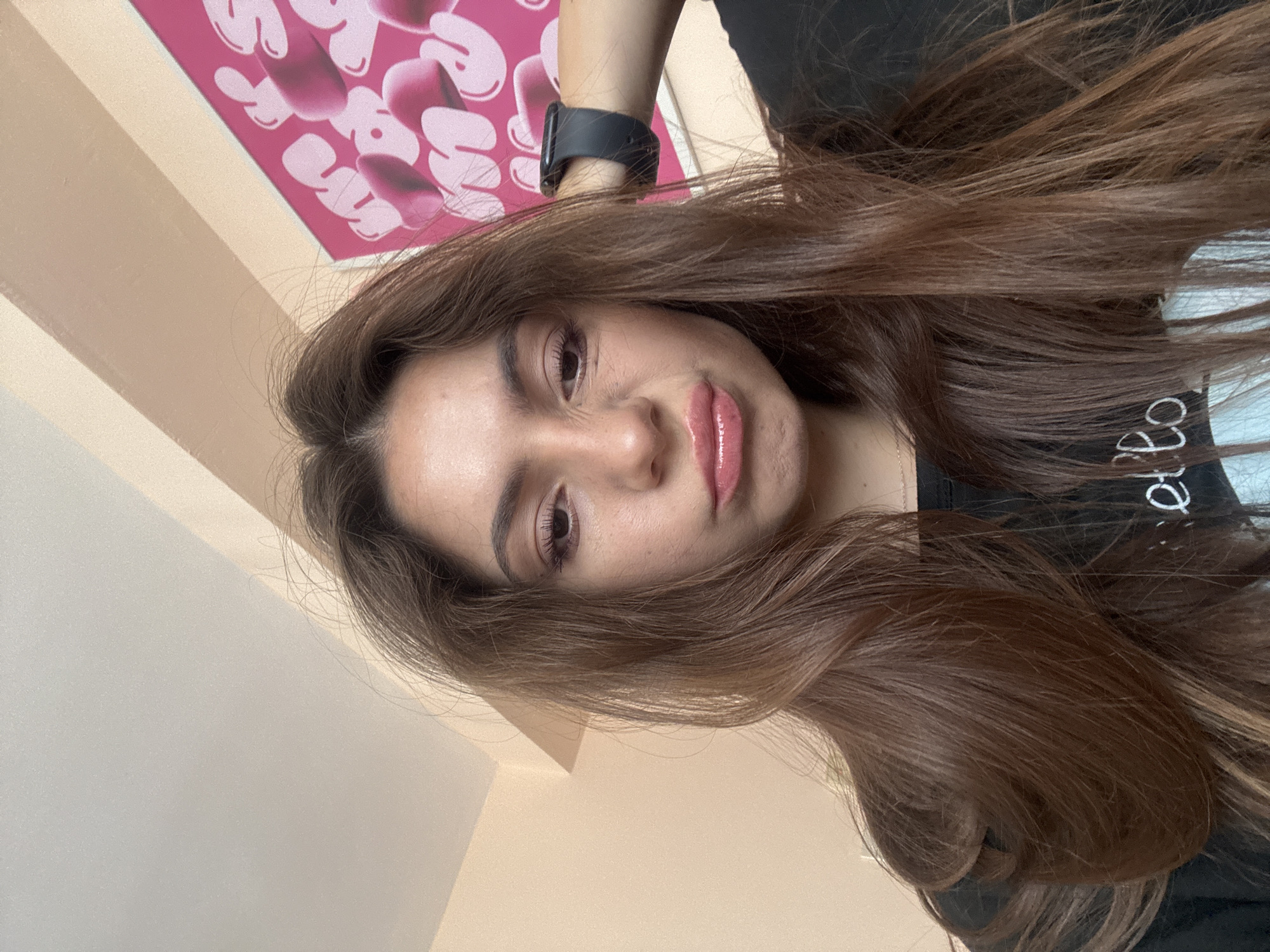 The media file is a close-up, sideways portrait of a young woman with long, brown hair. She is looking towards the camera with a slight smile. Her hair is prominent in the frame, falling around her face and shoulders. She is wearing a black top with some white lettering visible on the shoulder area. On her left wrist, she is wearing a dark-colored smartwatch.
The background consists of a wall painted in a warm, peachy-beige color, with diagonal lines suggesting a corner or architectural feature. In the upper left corner, a bright pink poster with abstract white and pink shapes is partially visible, adding a pop of color to the scene.
The lighting appears to be natural, coming from the side and above, casting soft shadows and highlighting her features. The overall mood is casual and relaxed. While the exact time of day and weather are not discernible, the lighting suggests it could be daytime. The presence of a poster and what appears to be an indoor setting indicates she is likely at home or in a similar personal space in Varna, Bulgaria. No other specific activities or events are evident beyond the posed portrait.