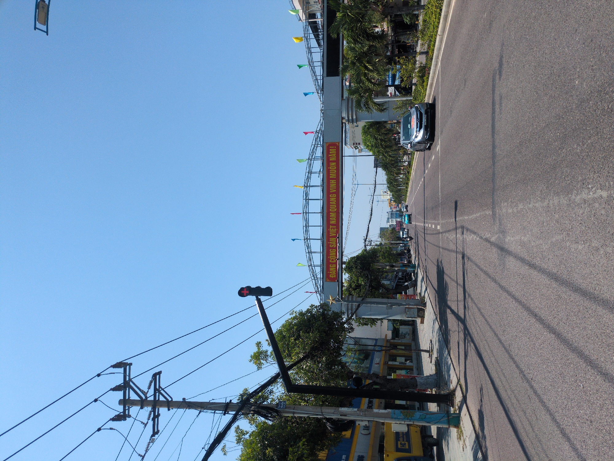 The image depicts a street scene in South Quy Nhon Ward, Vietnam. An asphalt road stretches into the distance, flanked by sidewalks and buildings. A car is present on the road. Overhead, a large metal archway decorated with colorful flags spans the road, bearing Vietnamese script. Power lines and poles run along the side of the road. A traffic light showing red is visible. Trees and vegetation are present along the roadside. The sky is clear and blue.