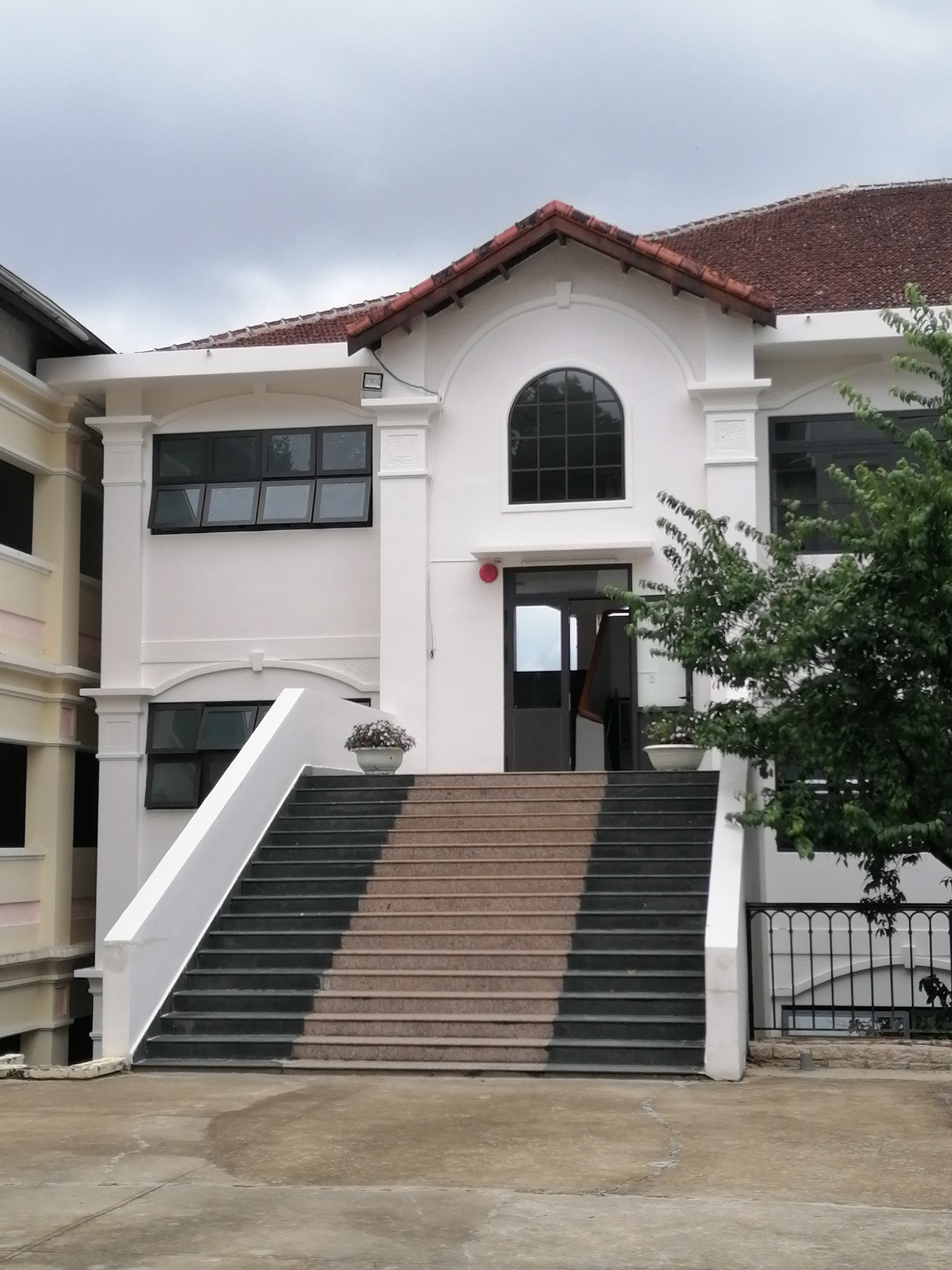 The image shows the exterior of a white building with a red tiled roof. A wide, grand staircase with dark gray and light brown steps leads up to the entrance, which features double doors. The building has multiple windows, including a large arched window above the entrance. To the left of the stairs, a portion of a light yellow building is visible. A lush green tree partially obscures the right side of the building. The scene appears to be in a well-maintained outdoor area, possibly a public space or institution, given the architectural style and the presence of a decorative potted plant on the staircase landing.