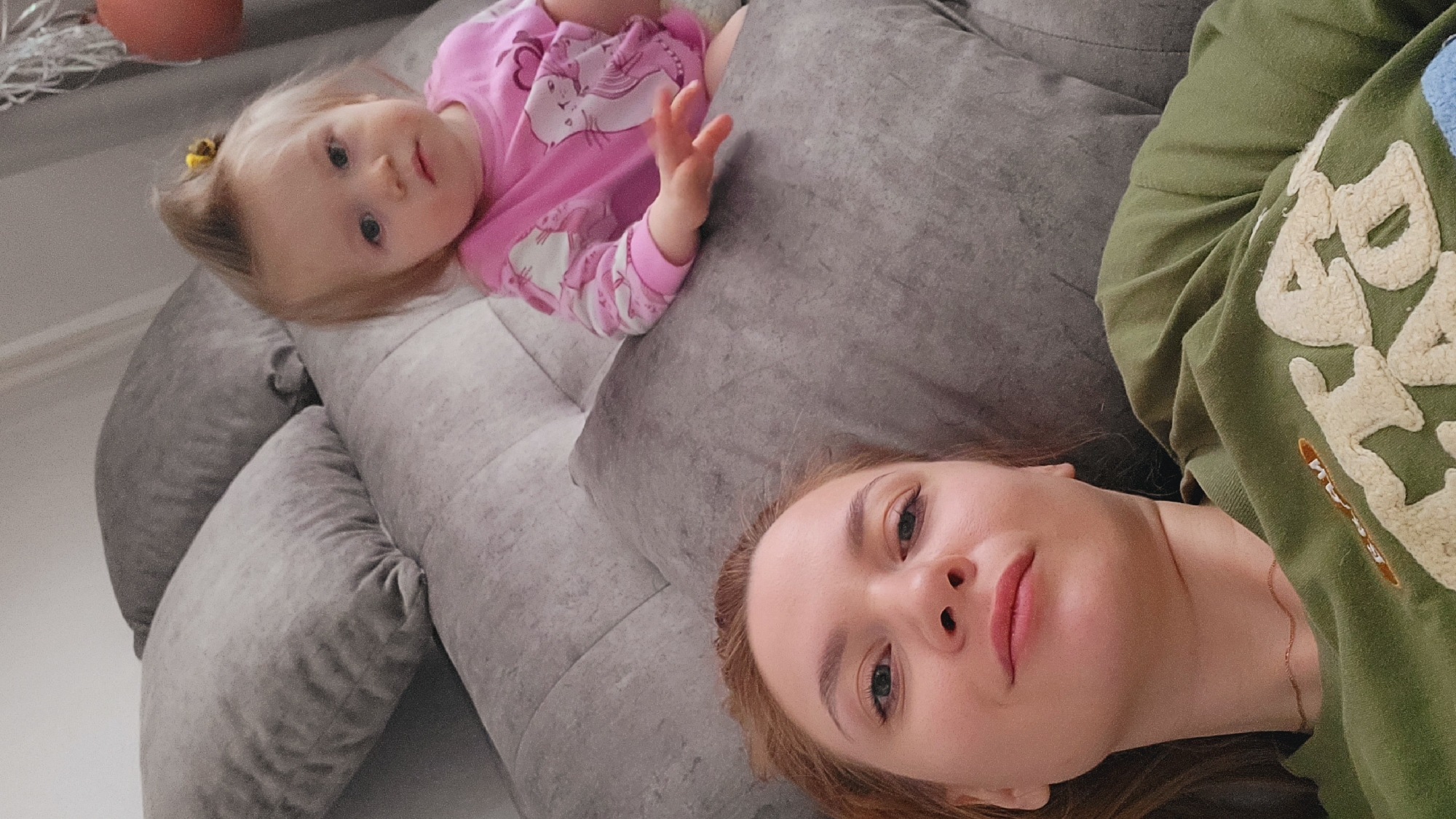 The image shows a woman and a young child lying on a gray sofa. The woman is lying on her side, facing the camera. She has fair skin, reddish-brown hair, and is wearing an olive green sweatshirt. A thin gold necklace is visible. The child is lying on their back next to the woman. They are wearing a pink long-sleeved shirt with a unicorn graphic. The child's hair is light blonde, and there is a small yellow hair clip on the side of their head. They have one arm raised, with their fingers extended. The sofa is gray and made of soft material. A potted plant sits on a ledge in the upper left corner of the image. The image suggests an indoor setting, likely a home. There is no information to confirm this image was taken in Chaikovsky, Russia.