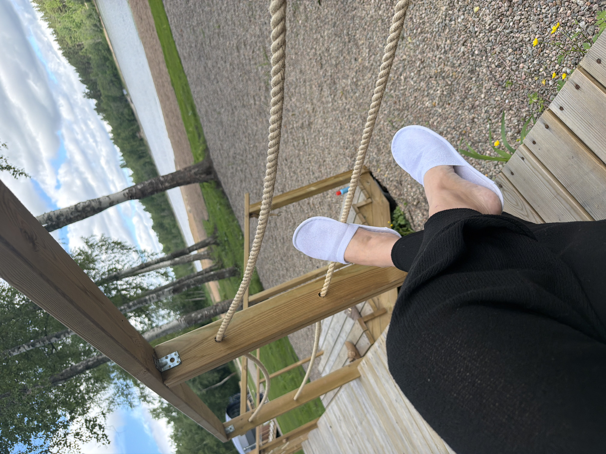 The image depicts a first-person perspective of someone resting on a wooden deck or boardwalk, wearing black pants and white slippers. The deck is situated outdoors, overlooking a body of water, likely a lake, with a sandy beach and a forested shoreline in the background. Birch trees are visible on the left side of the frame, their white trunks contrasting with the greenery. The sky is partly cloudy. The ground surrounding the deck is covered in gravel. A rope railing, made of twisted fibers, runs along the edge of the deck. The overall impression is one of a relaxed, natural setting, possibly a resort or recreational area.