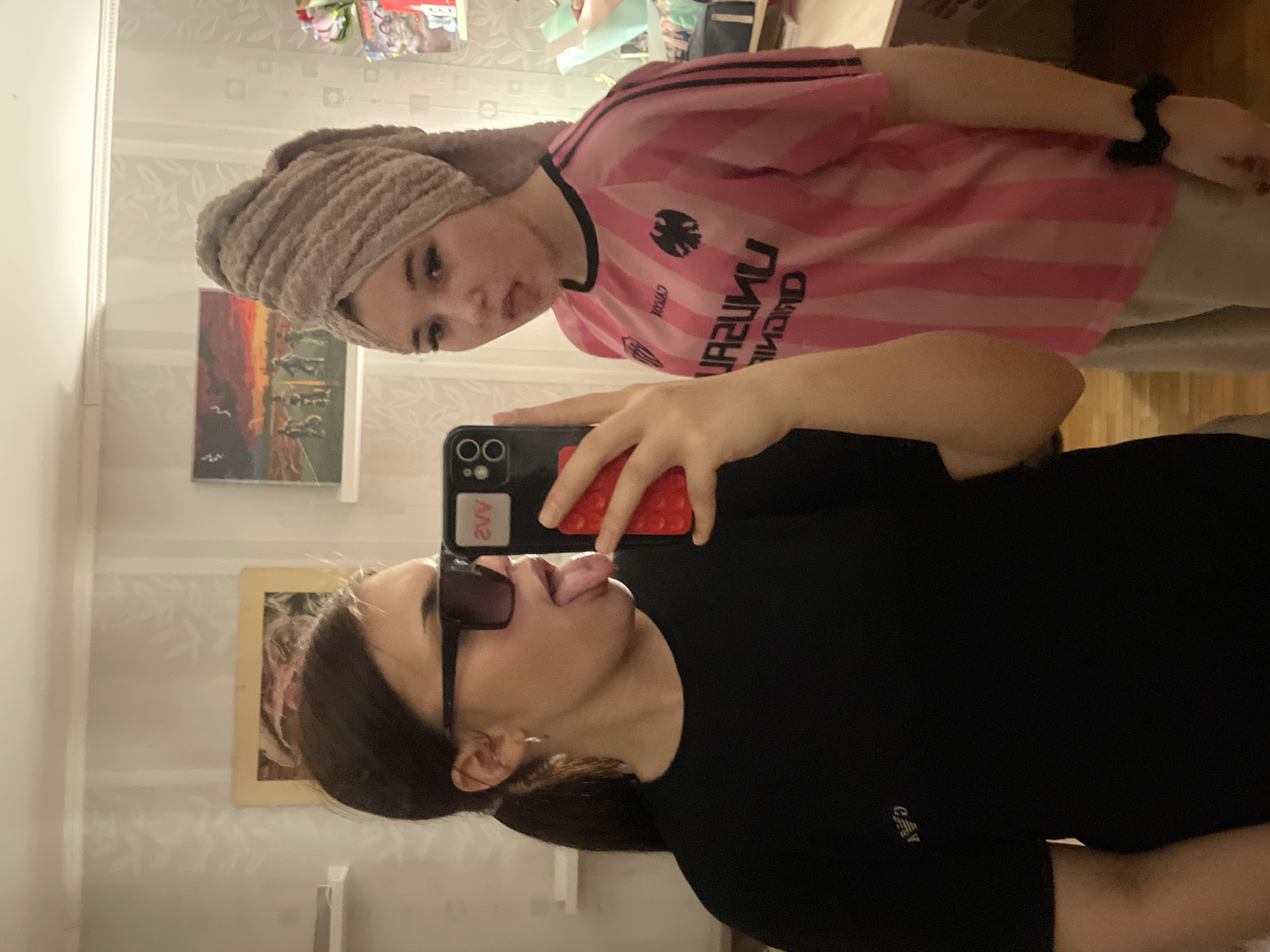 The image, taken in Vsevolozhsk, Russia, is a mirror selfie featuring two young women in an indoor setting. The photo is displayed on its side, with the subjects appearing horizontally.
In the foreground, a woman with dark hair, wearing a black t-shirt and dark rectangular sunglasses, takes the selfie. She holds a black smartphone with a textured red case, displaying "2VV" on its back. She is playfully sticking her tongue out. A small white "C&A" logo is visible on her t-shirt.
Reflected in the mirror behind her, a second young woman is visible. She wears a pink and black striped shirt and has a light-colored towel wrapped around her head, indicating she may have recently washed her hair. Text on her shirt, appearing reversed in the reflection, includes "UNREAL" and below it, another word that looks like "UNIOAD" or similar, along with a black bird-like emblem. She has a black scrunchie on her left wrist and looks directly towards the camera with a neutral expression.
The background reveals a room with light-colored wallpaper featuring a subtle leaf or floral pattern. A rectangular painting depicting a sunset landscape with distant figures and birds hangs on the wall. Other household items and shelves are faintly discernible in the deeper background. The scene is illuminated by indoor lighting, suggesting an indeterminate time of day.