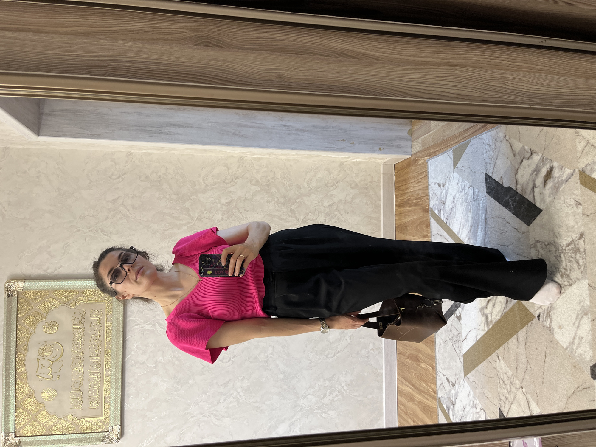 A woman with dark hair, wearing glasses, a bright pink shirt, and black pants is standing in what appears to be a hallway or room with a marble-patterned floor. She is holding a smartphone, seemingly taking a picture of herself in a mirror. A brown handbag is visible near her feet. On the wall to her left is a framed decorative artwork with Arabic calligraphy. The location is Kaspiysk, Russia.