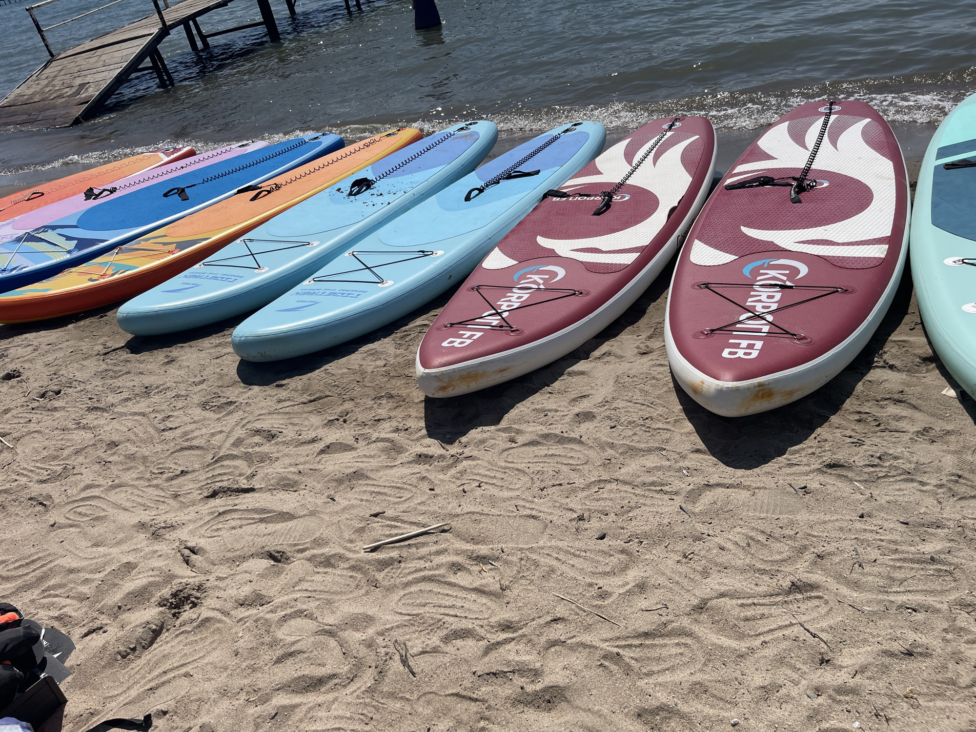 A close-up, eye-level, medium shot captures a row of stand-up paddleboards resting on a sandy beach. The boards are arranged in a line, with their tails facing the viewer and their noses pointing towards the water. The sand is a light brown, with visible footprints and debris scattered across its surface. In the background, the calm, rippling water of a lake stretches towards a wooden pier. The lighting suggests a sunny day, casting shadows on the sand. The scene appears to be set on the shore of Lake Issyk-Kul, near the village of Ornek in Kyrgyzstan.
