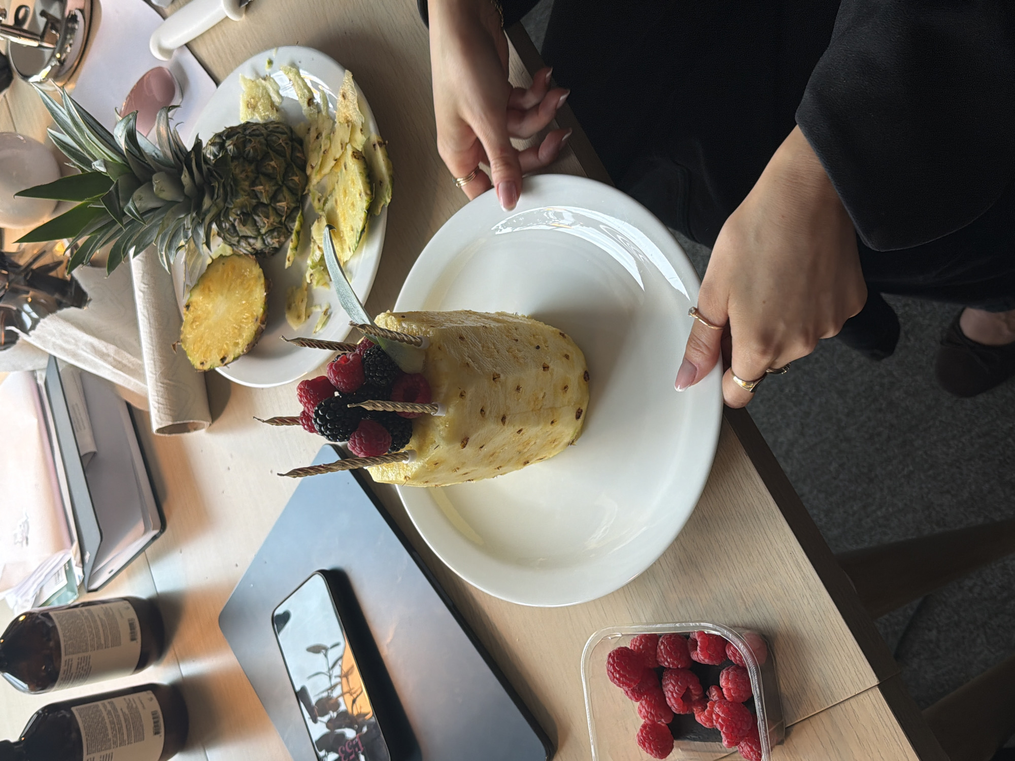 The image presents an overhead view of a light-colored wooden table in an indoor setting, consistent with a location like London, where food is being prepared or presented.
The central subject is a person's hands, adorned with rings and manicured nails, holding a white plate. On this plate, a largely peeled pineapple is artfully decorated with fresh raspberries and blackberries, which are attached using small decorative skewers or candles. This suggests a festive or celebratory fruit dish.
To the left, on a separate white plate, lies another pineapple, half-cut with its green crown still attached, alongside some discarded pineapple peels. This indicates that the fruit has recently been, or is in the process of being, prepared. A clear plastic container filled with ripe red raspberries is visible on the lower right side of the table, suggesting more fresh fruit is available.
Other items on the table include two dark amber bottles with small, illegible white labels, a smartphone reflecting what appears to be an indoor environment with natural light (possibly a window or plant), a white ceramic knife, a roll of paper, and some stacked documents or notebooks. The person is wearing a long-sleeved black top or dress. The scene conveys a moment of careful and deliberate food preparation, focusing on fresh fruit. No discernible text beyond the illegible bottle labels is visible.