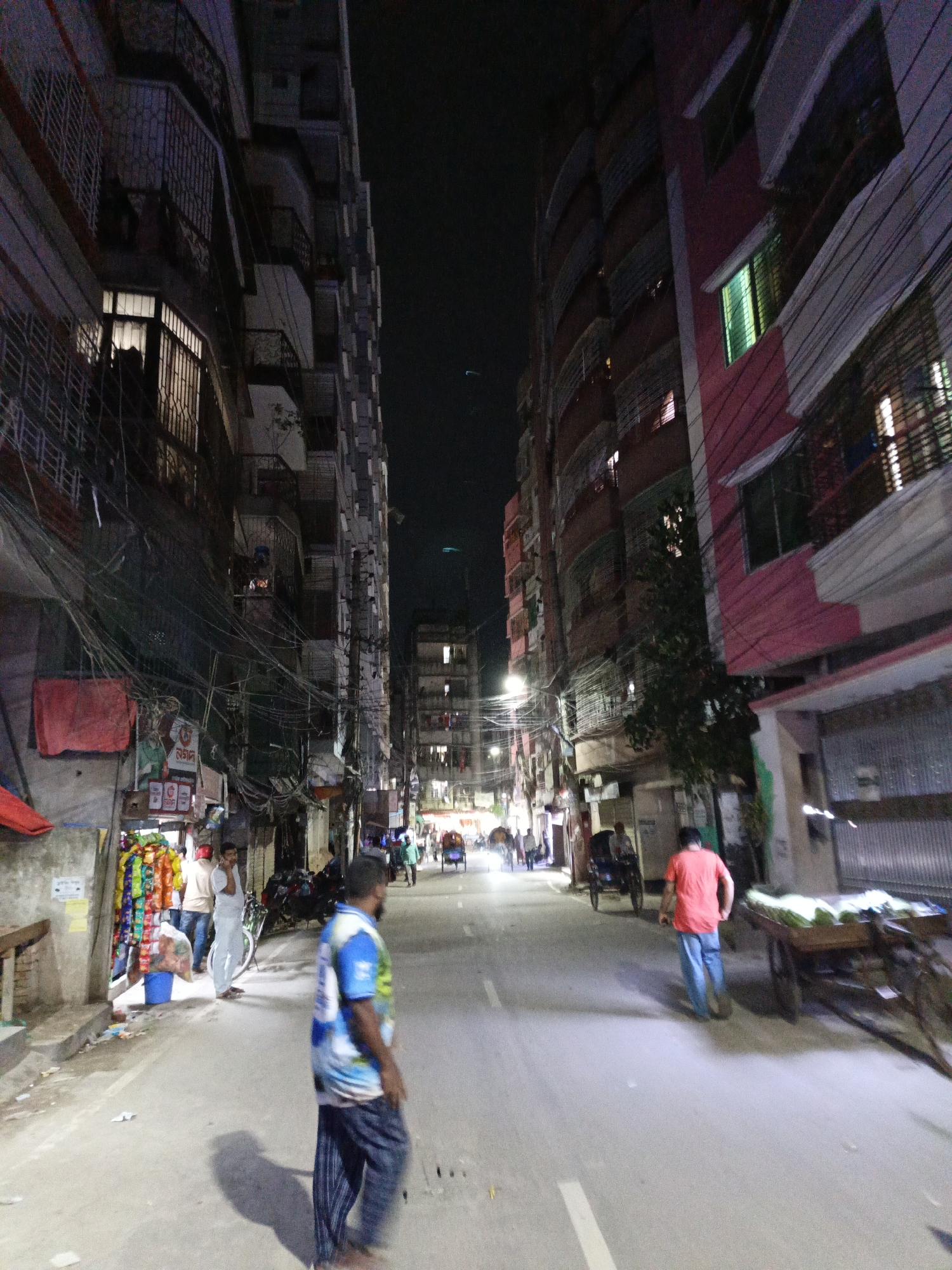 A street scene in Unknown, Unknown shows a narrow road lined with tall buildings at night. The buildings are a mix of colors, including red and grey, and have many windows, some with lights on. Power lines are strung haphazardly across the street. Several people are walking along the street, and there are carts and bicycles present. A man in a blue and white shirt walks in the foreground, while another man in a red shirt walks away from the camera further down the street. Streetlights illuminate the scene. There are shops and stalls along the side of the road.