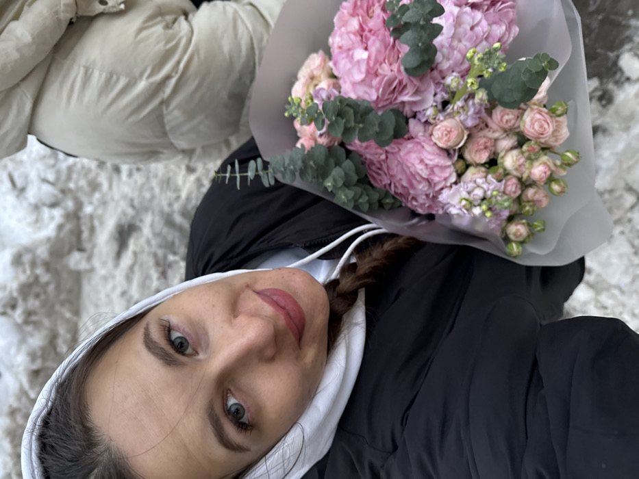 An outdoor image depicts a female subject holding a floral bouquet, partially obscuring a second individual, against a snow-covered background in Odintsovo, Russia. The primary subject, positioned in the lower-left to central frame, is a female with green eyes and a light brown braid visible under a white hood. She wears a black puffer jacket over a white hooded sweatshirt, the hood up, and looks directly at the camera with a subtle smile. Her lips are coated with reddish-pink lipstick. The bouquet, held in her left arm, features large pink hydrangeas, smaller pink spray roses, and green eucalyptus stems, wrapped in a light grey material. In the upper-right background, the torso and arm of a second individual are visible, clad in a light beige puffer jacket. The ground behind the subjects is covered with uneven, dirty white snow.