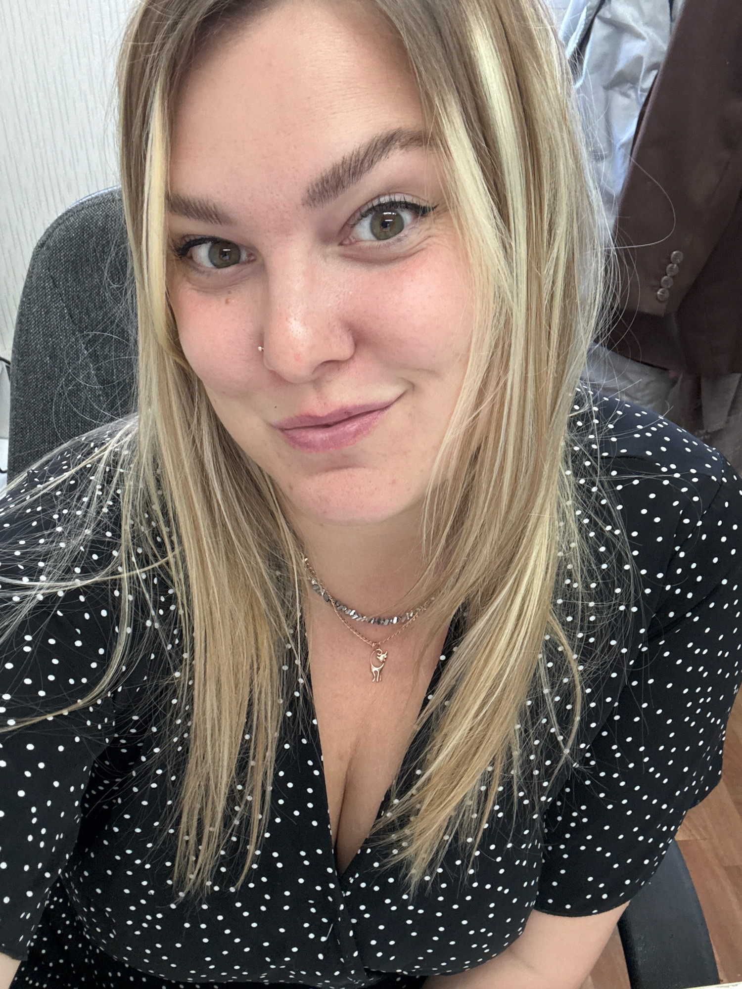 The image shows a close-up selfie of a young woman with blonde hair, green eyes, and fair skin. She is wearing a black and white polka dot dress and a necklace with a small pendant. Her head is slightly tilted, and she is smiling at the camera. She has a nose piercing. In the background, a gray chair, a brown suit jacket, and a textured wall are visible. The scene appears to be indoors, possibly an office environment, as indicated by the chair and suit jacket. There is no indication of the city of Noviy, Russia, or any specific location within it.
