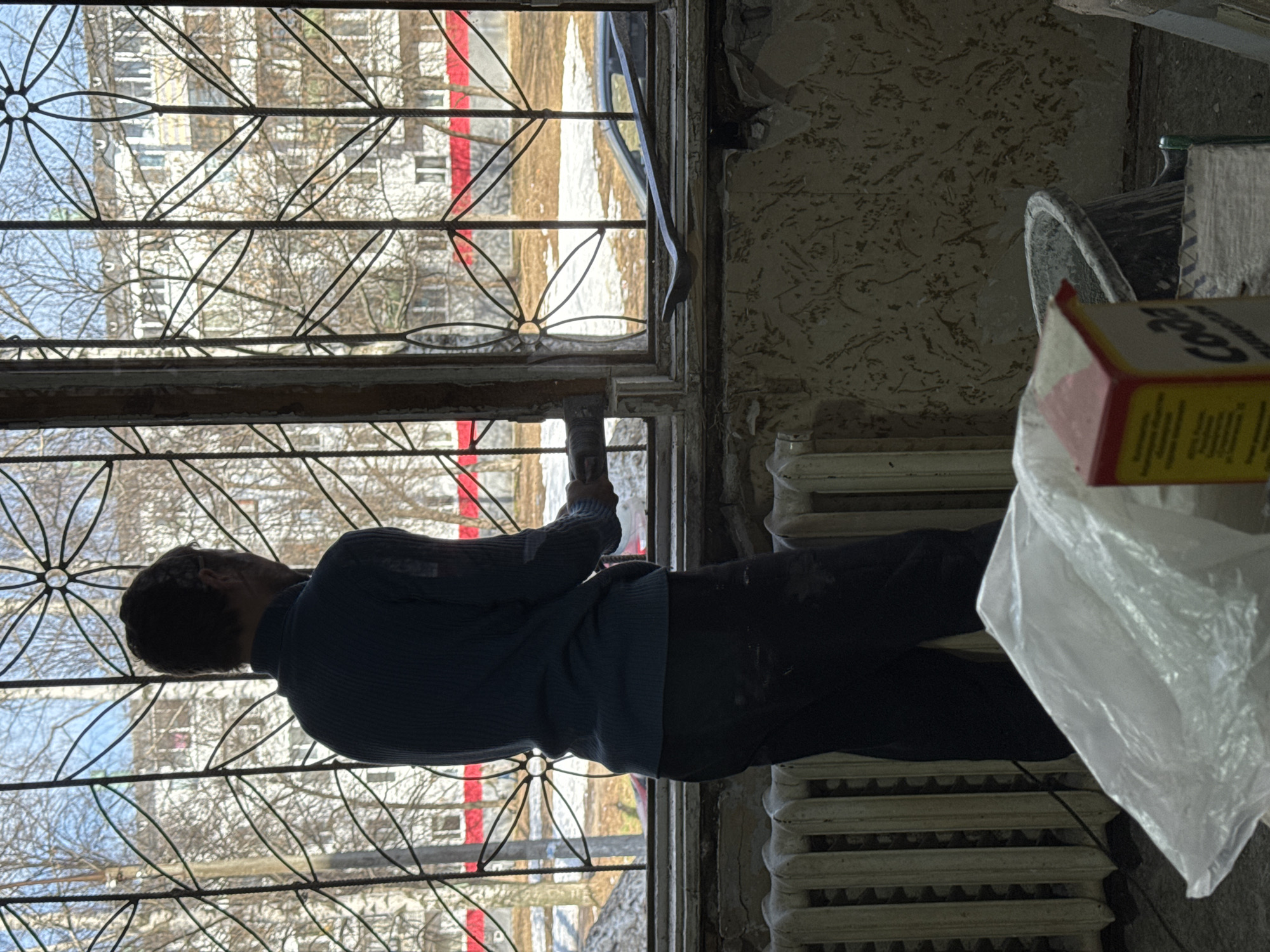 A person is working on a window in a room with peeling wallpaper and an old-fashioned radiator. The window has an intricate decorative ironwork pattern. Through the window, an apartment building with a red stripe on the facade is visible. Given the visual cues, this scene is likely located in an older building in Chaikovsky, Russia.