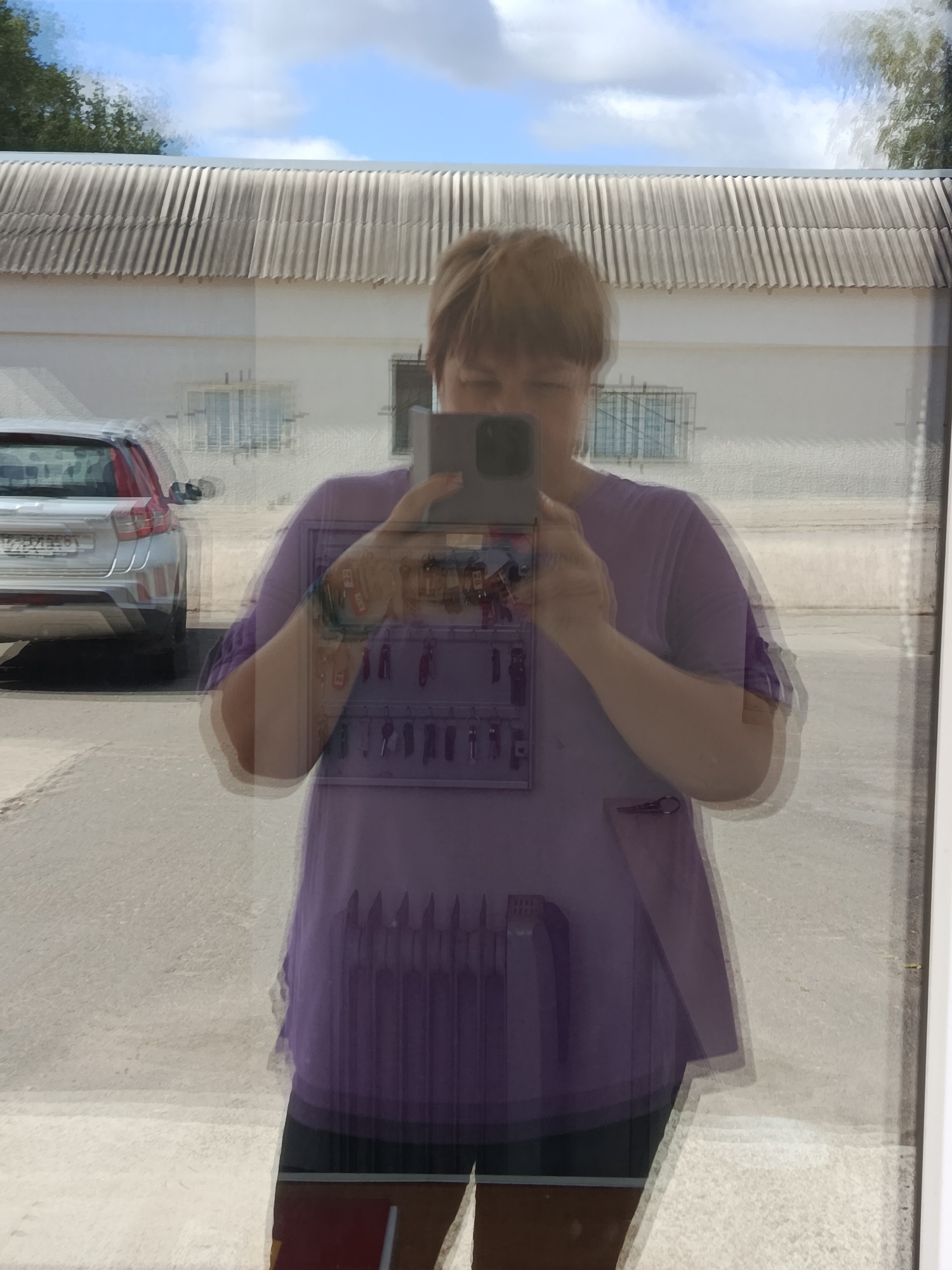 A person with short brown hair is reflected in a glass surface, holding a dark-colored smartphone to take a photo. They are wearing a purple t-shirt. Behind them, a silver car is parked, and further back, a light-colored building with a corrugated roof is visible. The reflection also shows a clear blue sky with some white clouds, and trees in the distance. The scene appears to be outdoors, possibly in a parking lot or near a building in Svietlahorsk, Belarus, given the context. A rack of keys is visible hanging on the wall behind the person in the reflection.