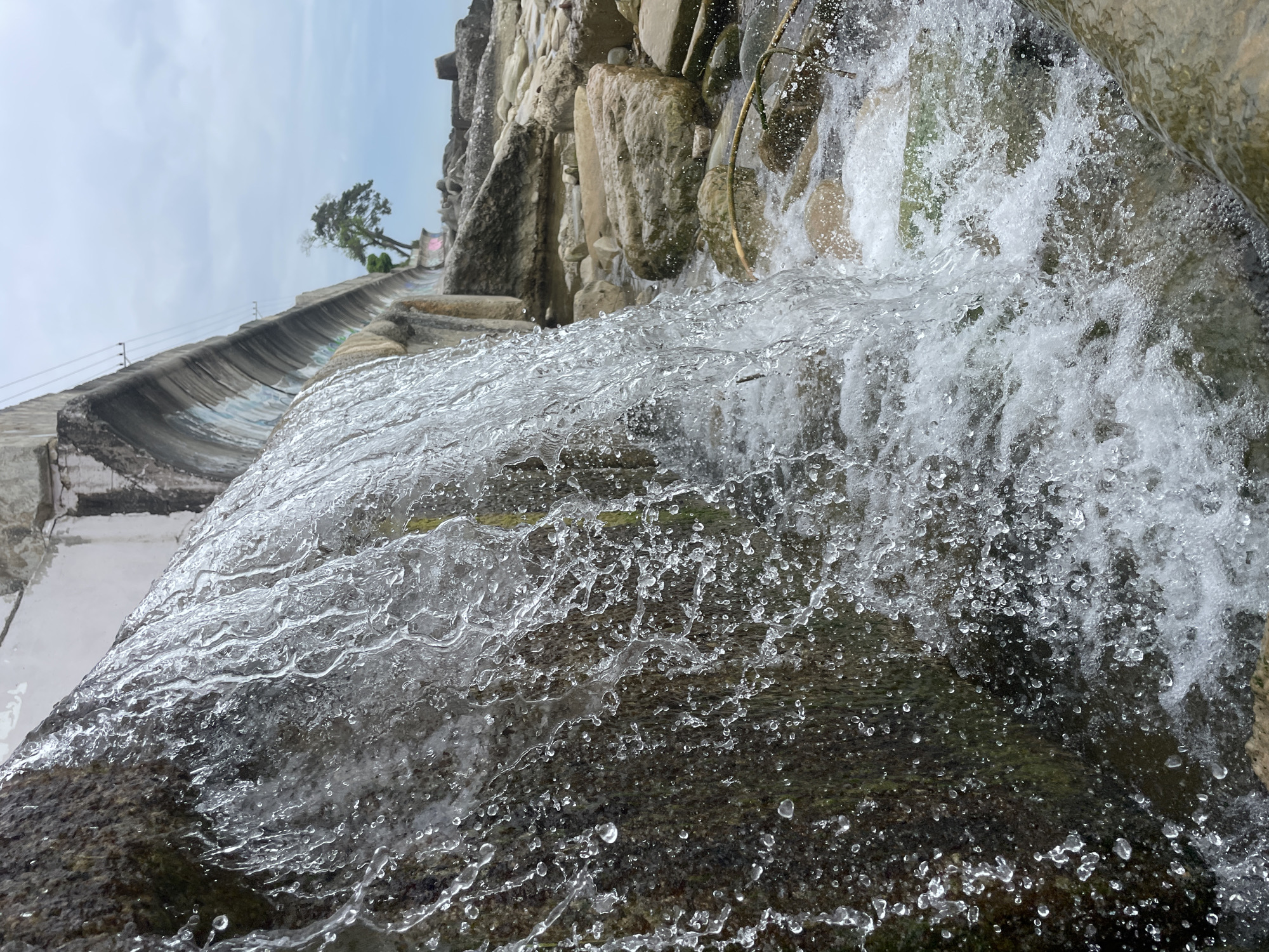 The image shows a waterfall cascading down a rocky structure. A concrete trough with graffiti runs along the left side of the image, leading to the waterfall. A small tree grows along the top of the trough. The sky is visible in the upper left corner. The water is shown in motion, with many droplets and splashes visible. Based on the style of architecture and landscape, this is likely located in Sochi, Russia.