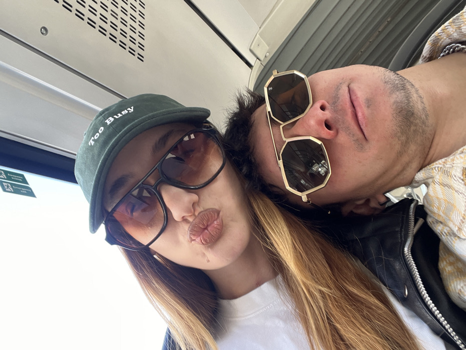 The image captures two individuals, one female and one male, posing for a selfie inside what appears to be a metro train in Sofia, Bulgaria. The female is positioned slightly above and behind the male, with long light brown hair draped over her shoulder. She wears a dark green baseball cap with "Too Busy" embroidered in white on the front, and large, dark gradient sunglasses. Her lips are pursed in a kiss-like gesture, and a small, healed scar is visible on her left cheek. She is wearing a white crew-neck garment. The male, in the foreground, has dark hair and stubble on his face. He wears gold-framed, hexagonal-shaped sunglasses with dark lenses, a light-colored plaid shirt, and a black leather jacket. A silver chain is visible around his neck. The female's head is resting near the male's upper body, suggesting close proximity. The background shows the interior of a public transport vehicle with white walls and ceiling panels, overhead ventilation grilles, and a window frame visible on the left side. A green sign with white pictograms is visible on the ceiling panel above the female.