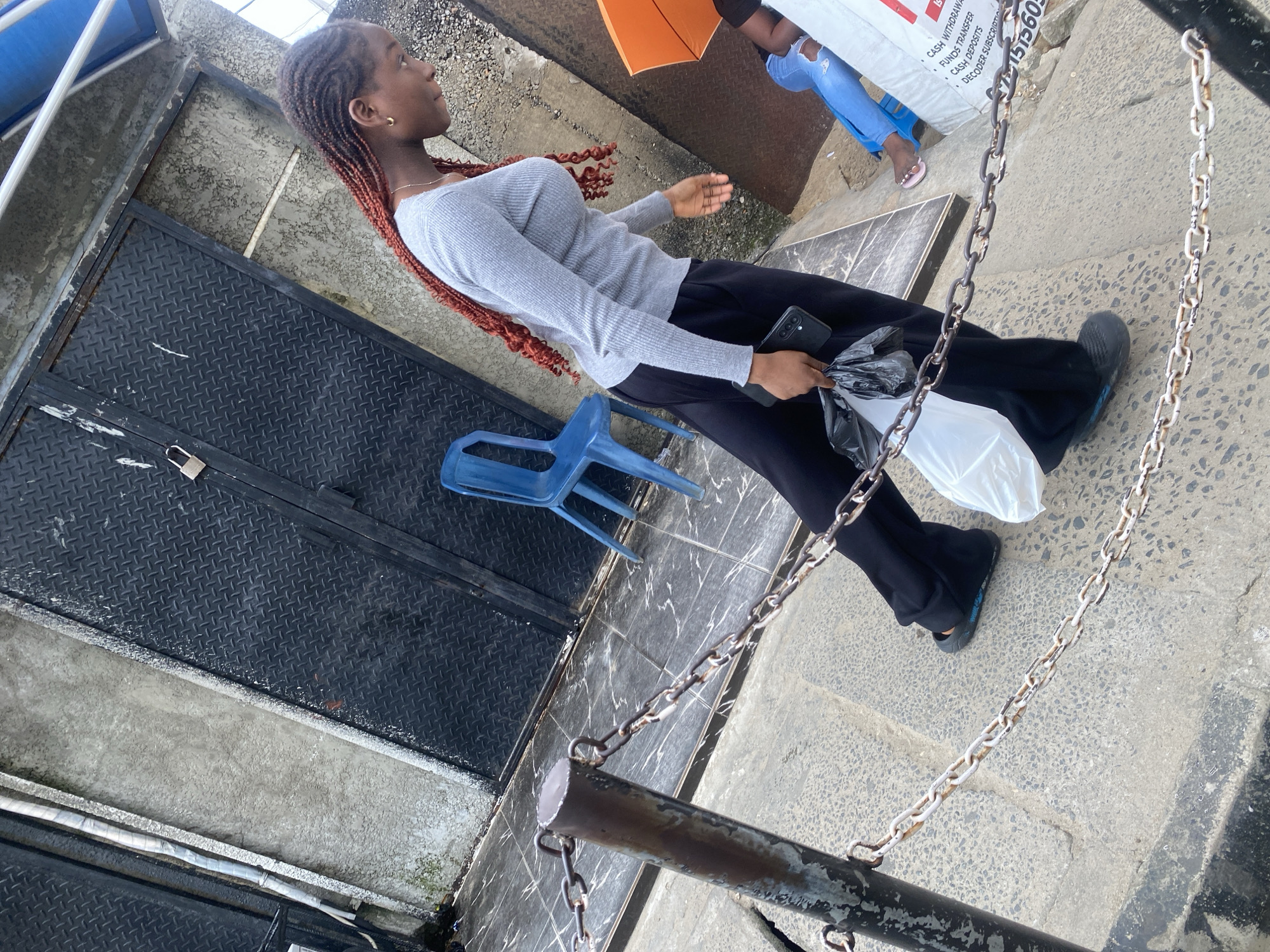 A young woman with braided, reddish-brown hair walks diagonally across the frame from right to left, viewed from a slightly elevated, angled perspective. She is wearing a light gray, long-sleeved, ribbed top and loose black pants. In her right hand, she holds a black smartphone, and in her left hand, she carries two plastic bags, one white and one black. She is wearing dark slip-on shoes.
In the background, a large, dark metal door with a diamond plate pattern is visible, secured by a padlock. To the left of the door is a rough concrete wall. A bright blue plastic chair is placed in front of the door, and another similar chair is seen further to the right, partially obscured by the woman.
A rusty metal post with a chain running across it is in the foreground, extending from the bottom left corner towards the center of the image. The ground is paved with gray and white marbled tiles. To the far right of the frame, another person is visible, wearing blue jeans and pink flip-flops, seated on a blue chair and holding an orange umbrella. A white bag with text, including "CASH WITHDRAWAL" and "FUNDS TRANSFER", is next to them. The overall environment appears to be an outdoor urban setting.