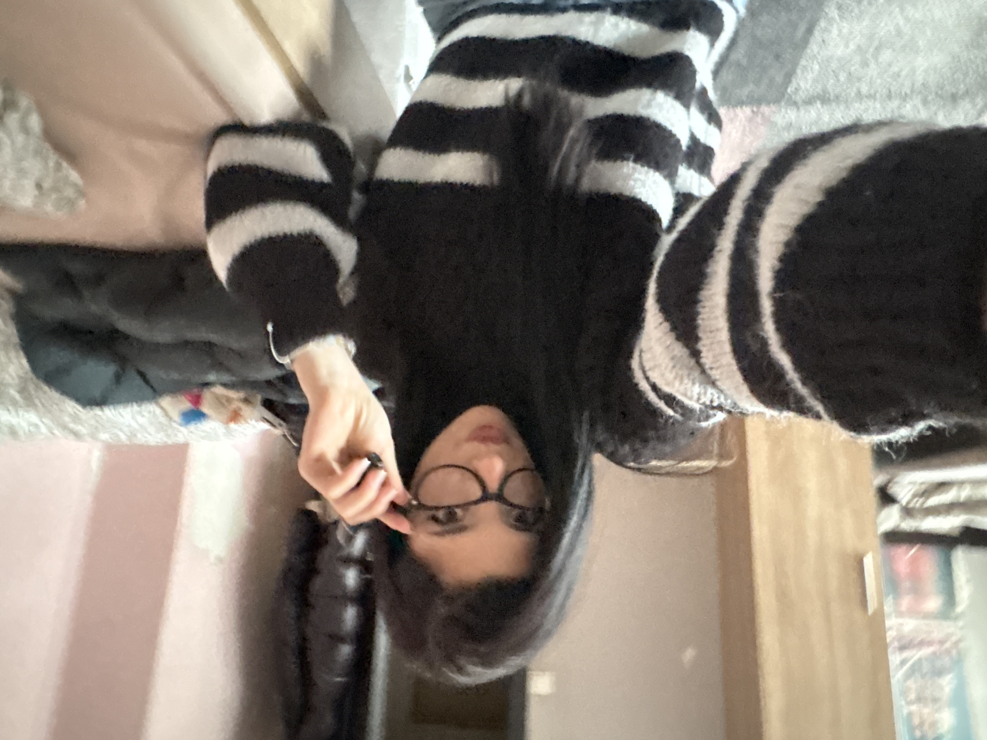 A person with black hair and glasses is wearing a black and white striped sweater. The person is holding their glasses. A black jacket is visible to the left. A bed is in the background. A wooden door or cabinet is visible to the right. The exact location within Sofia, Bulgaria, cannot be determined from the image.