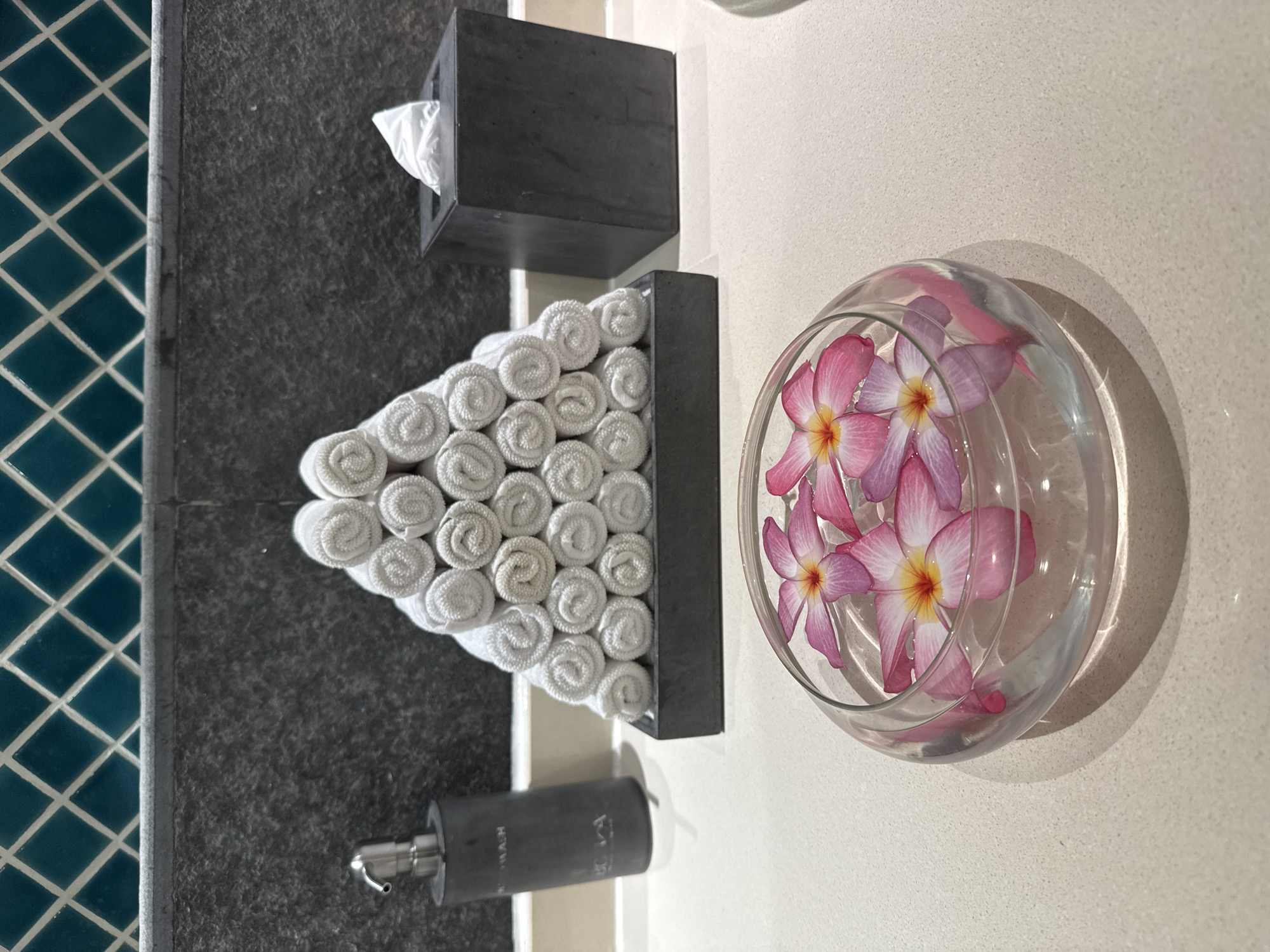 A still-life image shows a bathroom counter with decorative elements. In the foreground, a glass bowl filled with clear water holds several pink and yellow plumeria flowers. Behind the bowl, a dark gray stone tray displays a pyramid of neatly rolled white towels. To the left of the towels, a dark gray soap dispenser with a silver pump is positioned. To the right of the towels, a dark gray tissue box with a white tissue peeking out sits on the counter. The background features a dark gray textured wall and a wall adorned with teal diamond-shaped tiles. The image is set in a bathroom, likely in a hotel or spa, in Bac Nha Trang Ward, Vietnam.