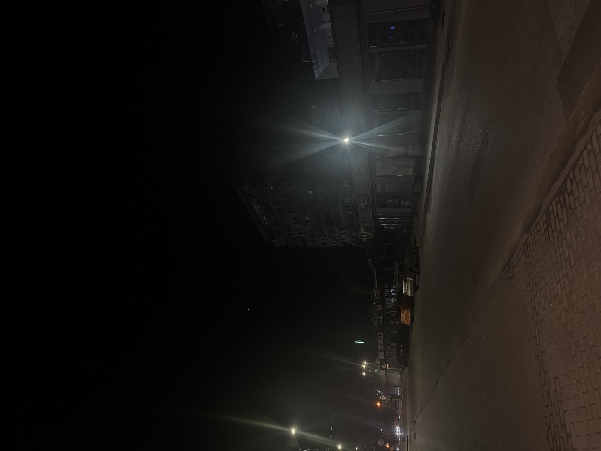 This is a nighttime street scene in Dnipro, Ukraine, likely on a commercial street. A multi-story building with many windows dominates the right side of the frame, its facade illuminated by a bright, starburst-like light source, possibly a streetlamp. Below the building, a row of storefronts with large glass windows lines the street. Some of these windows are covered with dark sheeting, suggesting the businesses are closed. A few cars are parked along the street, including an orange sedan and a white sedan. The street itself is paved, with a brick sidewalk visible on the left side, partially illuminated by streetlights. The sky is dark, with a faint celestial object visible. The overall impression is one of quietude and emptiness, typical of a city street after dark.