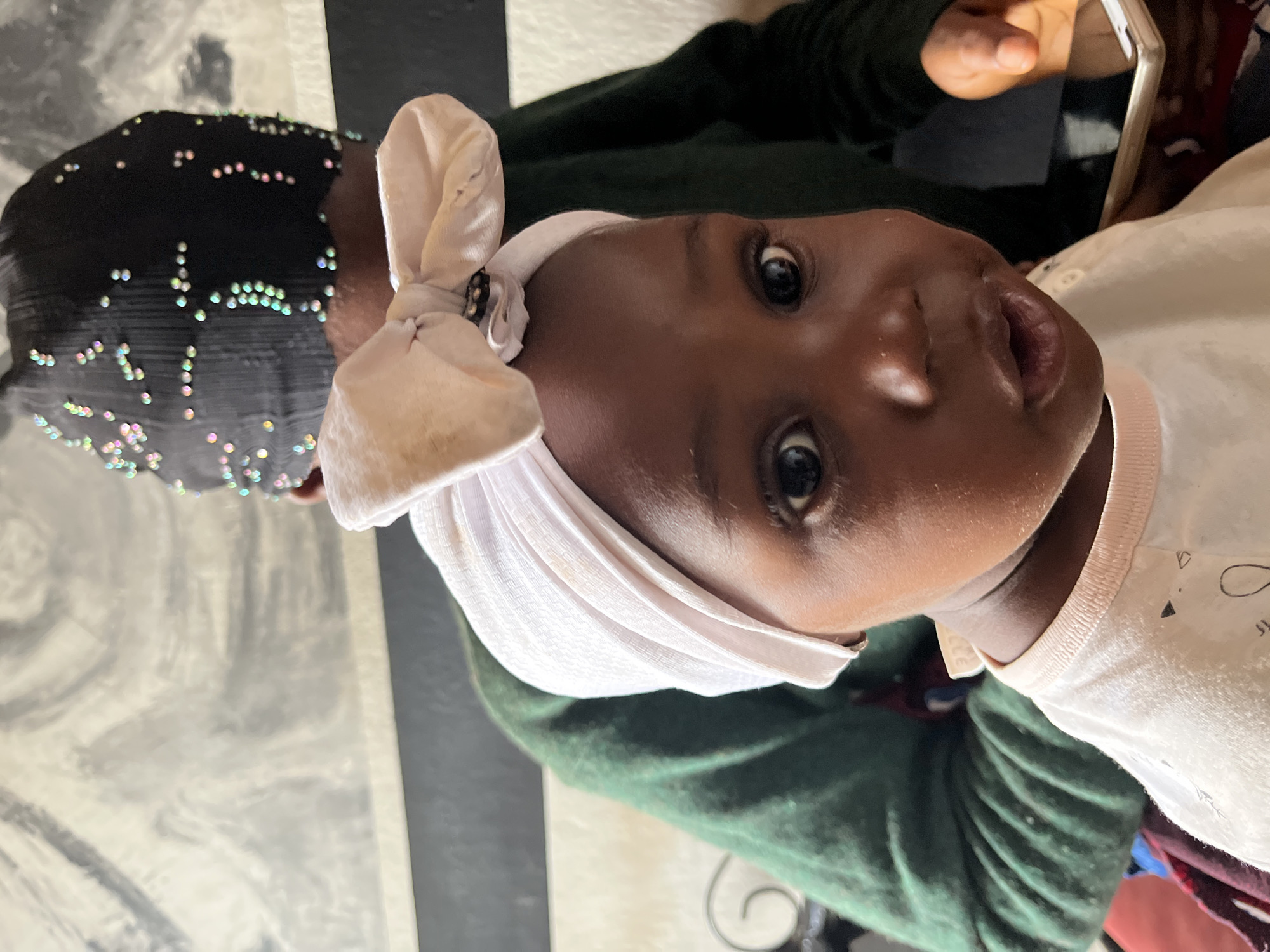 The image captures a close-up of a young baby looking directly into the camera. The baby has dark skin, wide, dark eyes, and a slightly open mouth, conveying an expression of curiosity or attentiveness. A light pink, soft fabric headwrap is tied into a bow on top of the baby's head. They are wearing a light-colored, possibly white or off-white, top with faint, dark abstract patterns.
Partially visible in the background is an adult, mainly their head and upper body. The adult is wearing a dark head covering, likely black, adorned with numerous small, iridescent beads or sequins that reflect various pastel colors like pink, green, and blue. They are dressed in a dark green long-sleeved garment. The adult's right hand is visible, holding a white mobile phone.
The setting appears to be indoors, with a floor or table surface visible in the background featuring a black and light-toned, possibly grey or white, striped pattern with some textural variations. The lighting is soft and consistent with an indoor environment. There is no visible text in the image. The location is Barakin Lerawi, Nigeria.