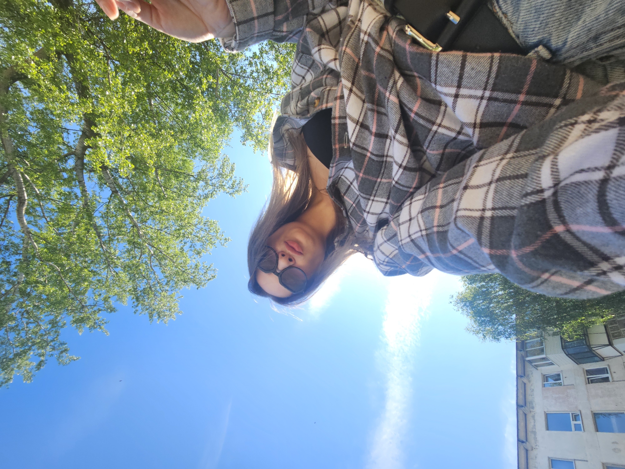 A woman is taking a selfie from a low angle, looking down at the camera. She is wearing sunglasses and a gray and pink plaid shirt. In the background, there is a clear blue sky with a few trees visible. A building is also partially visible in the background. The image was taken outside of a building located at Lenina Street, 16, Chaikovsky, Russia.