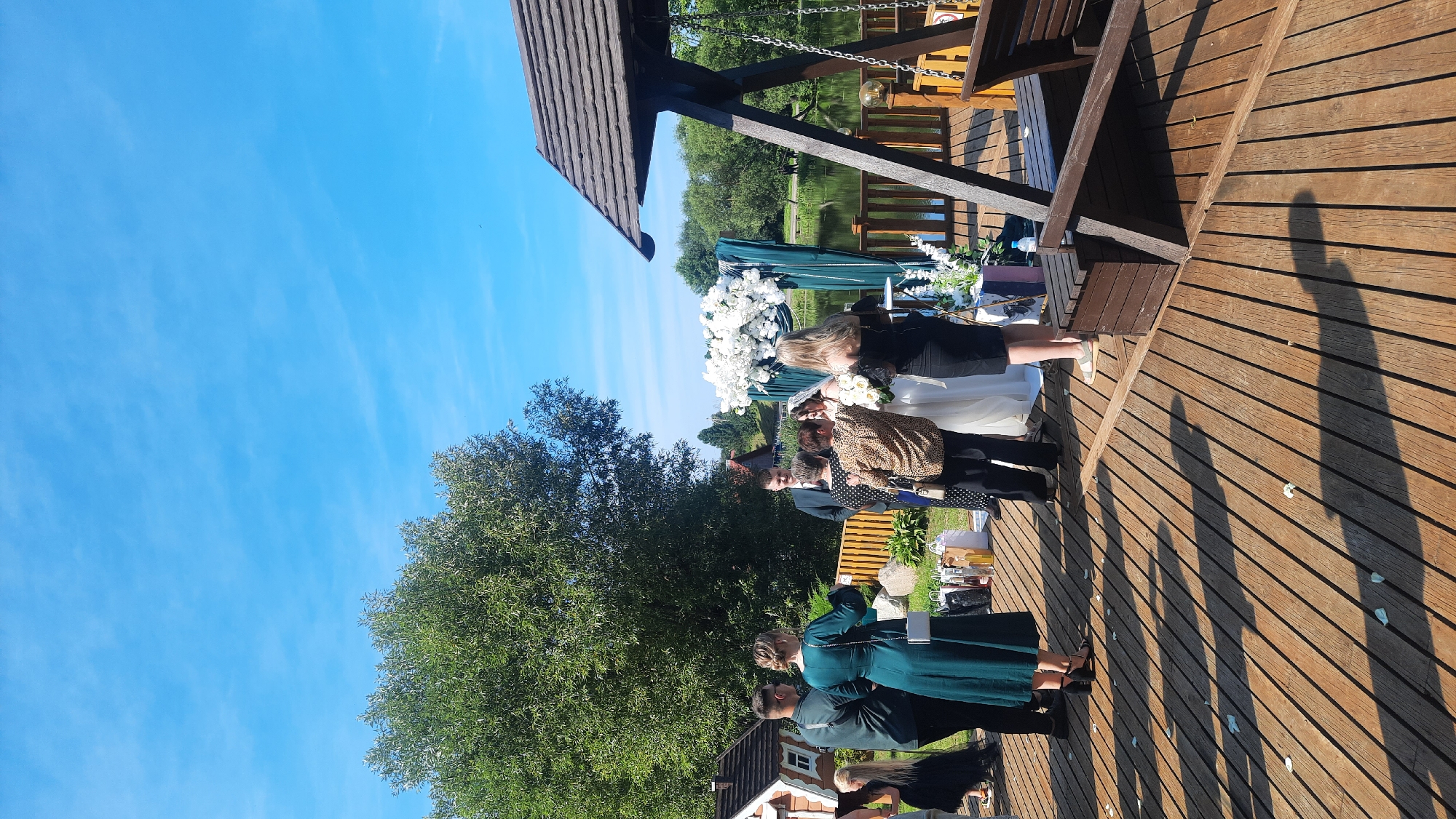 A wedding ceremony is taking place outdoors on a wooden deck next to a body of water, under a clear blue sky with wispy clouds. A bride in a white wedding dress and a groom in a dark suit stand at an altar adorned with white flowers. Several guests are gathered around them, some dressed in formal attire. To the right of the frame, a wooden swing set with two benches is partially visible. In the background, lush green trees and residential buildings are present. The scene is set in the agricultural municipality of Zhodzishkawski Sell' sovi in Belarus.