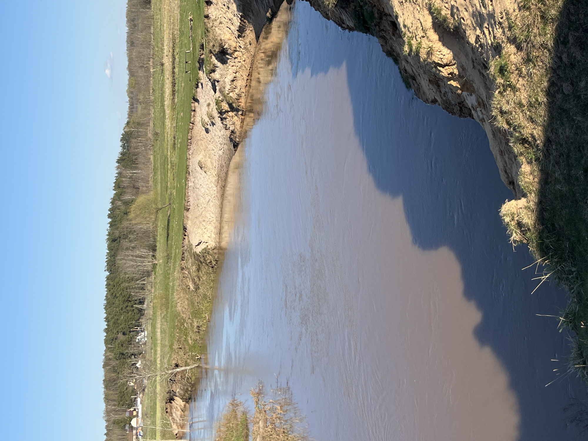 The image depicts a muddy river flowing through a steep, eroded bank in Razdolye, Russia. On one side, a tree-lined field borders the river's edge. The other side has a high, unstable cliff face, sparsely covered with grass. The water reflects the sky, adding a muted blue tone to its brown hue. The sun casts long shadows along the riverbank, enhancing the contrast of the scene. Some buildings are seen in the distance.