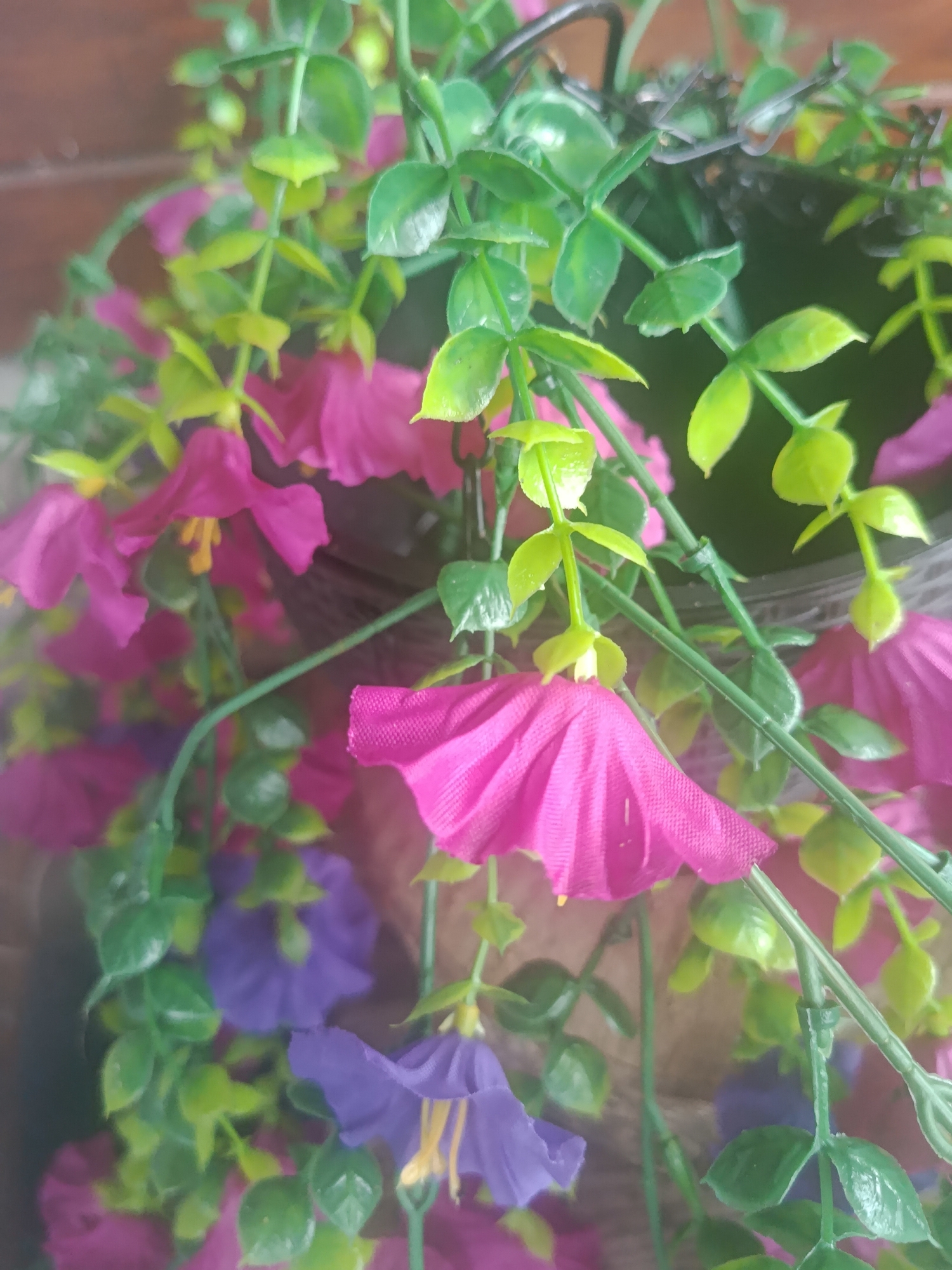 The image shows a close-up of artificial flowers, primarily pink and purple, interspersed with green artificial leaves and stems. The flowers are hanging, suggesting they are part of a planter or garland. The background is blurry, with what appears to be a wooden surface on the left and a dark, possibly woven material on the right, which could be the planter itself. There are no people or animals visible. The scene is indoors or in a sheltered outdoor area, given the artificial nature of the plants and the lighting. The location in the city of Верея, Russia, is not discernible from the visual information provided in the image itself.