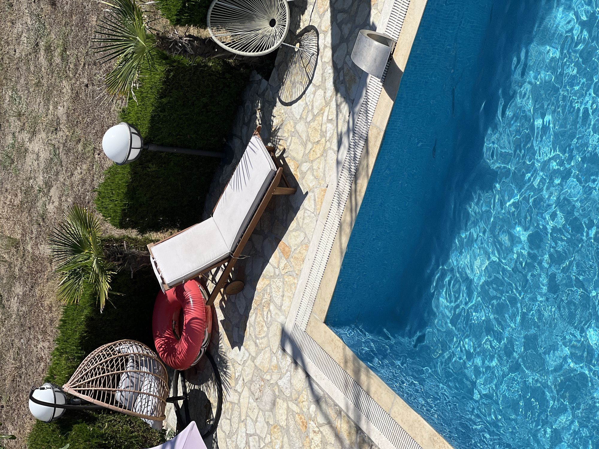 An aerial, high-angle, daylight, outdoor, full shot captures a serene poolside scene in Donji Štoj - Shtoji i Poshtëm, Montenegro. The azure blue water of a swimming pool fills the foreground, its surface rippling gently under the sunlight, with faint shadows of what appear to be fish visible beneath the surface. The pool's edge is bordered by a beige coping and a white, slatted drainage system. Surrounding the pool is a patio paved with irregular, light-colored flagstones.
Adjacent to the patio, a neatly trimmed, dark green hedge forms a boundary. To the left of the frame, a light pink, rounded, fabric-covered lounge chair or chaise longue is partially visible. Next to it, a wicker hanging egg chair with gray cushions sits on a stand. In front of the egg chair is a large, inflatable red ring.
Further into the scene, a wooden lounge chair with a white cushion is positioned on the patio, its wheels visible. To the right, a white, wire-frame chair with a circular design casts a distinct shadow on the flagstones. Two outdoor lamps with white spherical tops are placed near the hedge, one to the left and one to the right, both mounted on dark poles. A few palm trees with sparse fronds are interspersed among the hedges. The background consists of dry, dusty ground with sparse vegetation. The overall impression is a tranquil, sun-drenched recreational space.