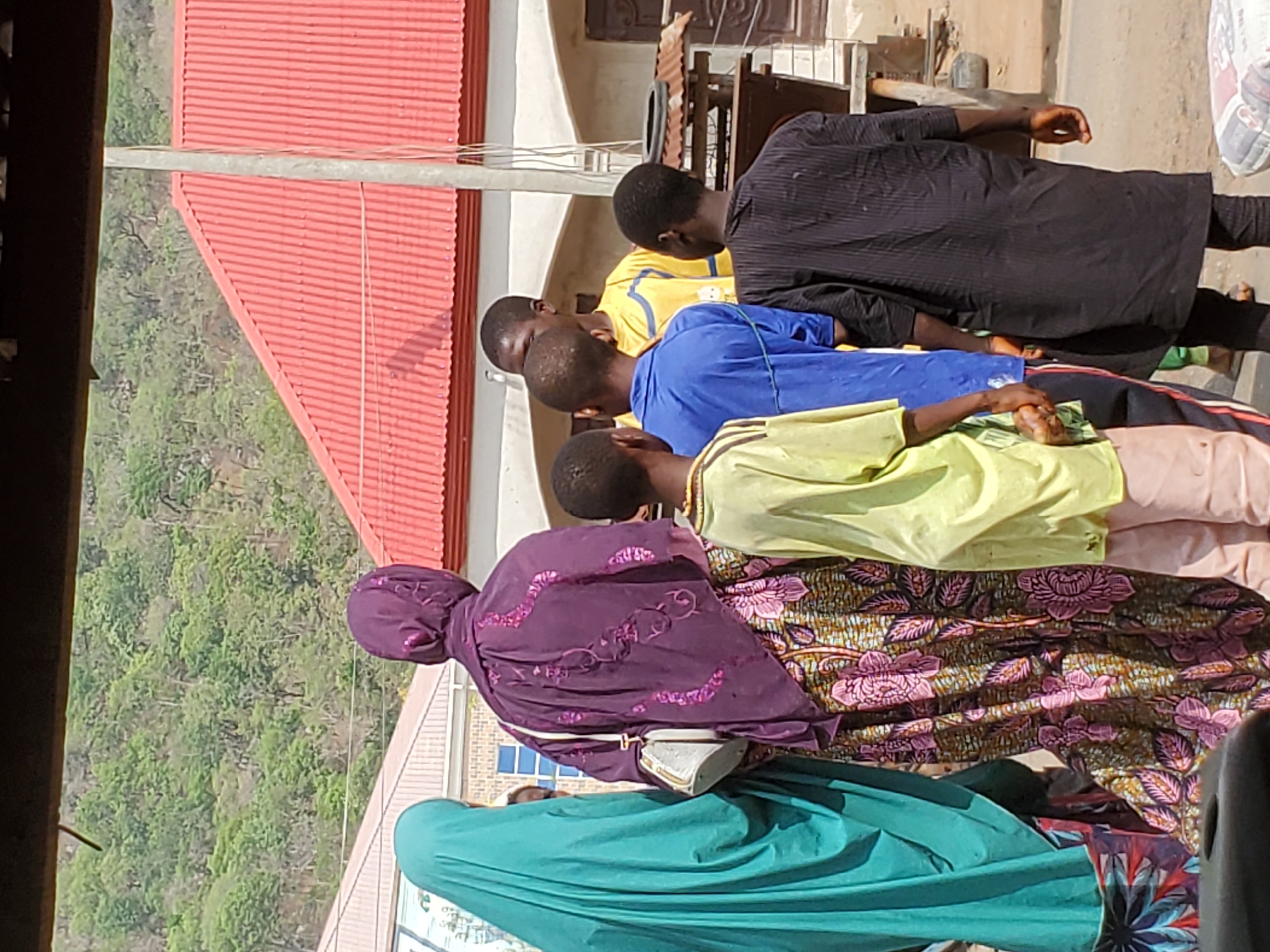 The image depicts a group of individuals standing outdoors. They appear to be of African descent. One is wearing a green headscarf and a floral print dress, and another a purple headscarf and a floral print dress. Several males are wearing shirts of various colors: black, yellow, blue, and light green. They stand in front of a building with a red roof, and a forested area is visible in the background. The location is identified as Mararaba, Nigeria.