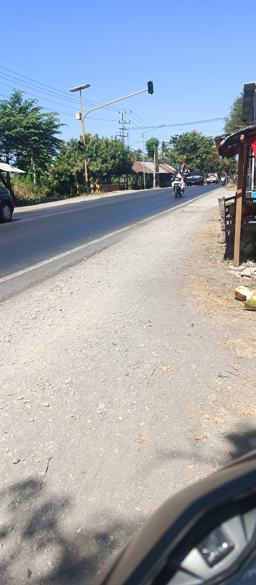 A sunny, clear day is depicted on a rural road in Jatisari, Indonesia. The road is paved with asphalt and has a gravel shoulder. A traffic light is visible on a pole with a solar panel. Several motorcycles and a car are traveling on the road. On the side of the road, there are trees, small buildings, and some coconuts. The foreground shows part of a vehicle, likely a car, with a side mirror reflecting the sky.