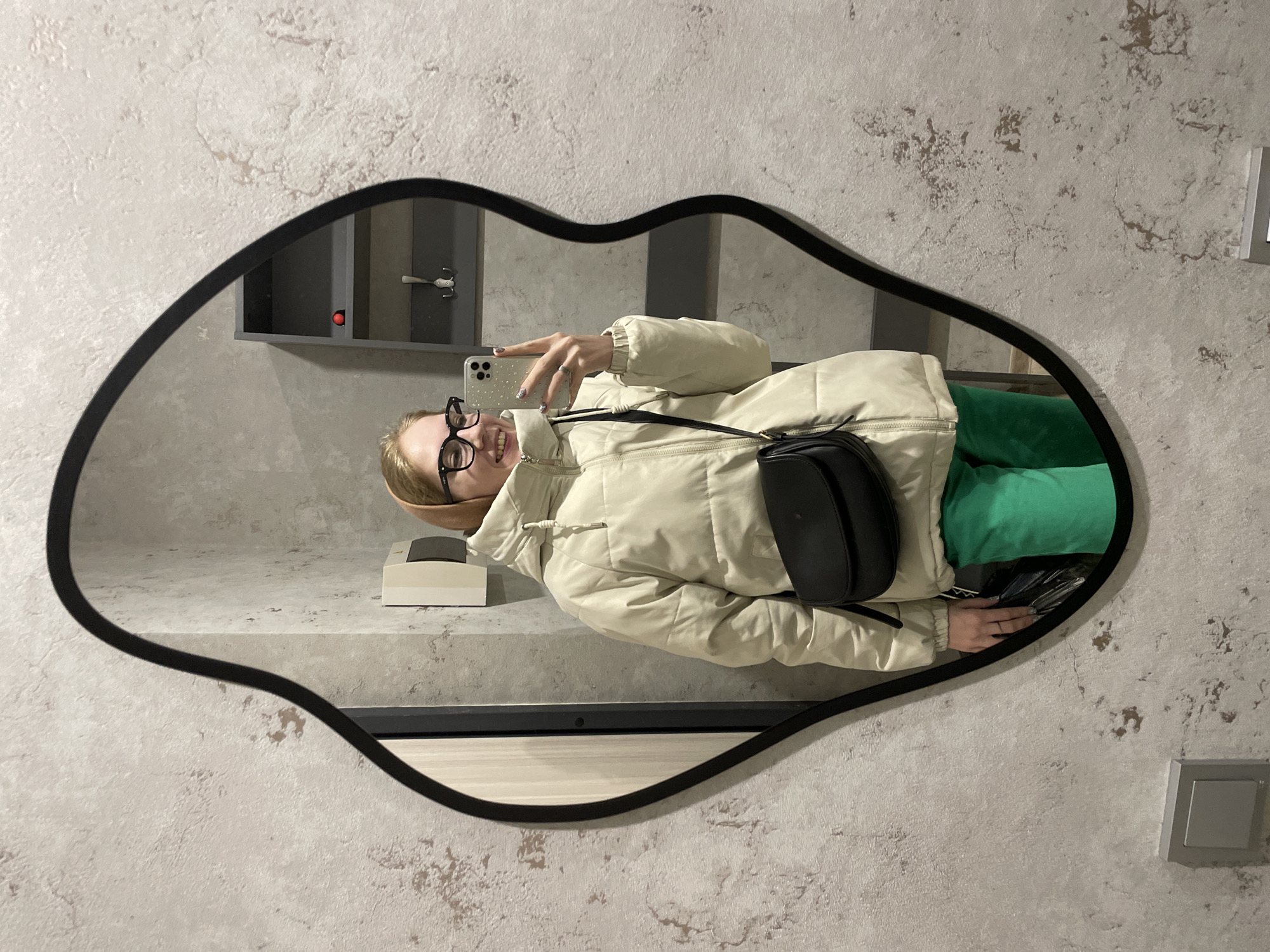 A woman is taking a selfie in a lip-shaped mirror. The mirror is in a bathroom with gray walls. The woman is wearing a light beige puffer jacket, green pants, and glasses. She has a black purse slung over her shoulder. A small box and some other items can be seen on a ledge or countertop in the reflection. The environment includes a coat hook, lightswitch, and what seems like a locker. It is not possible to verify the location in the city of Izhevsk, Russia from the given image.