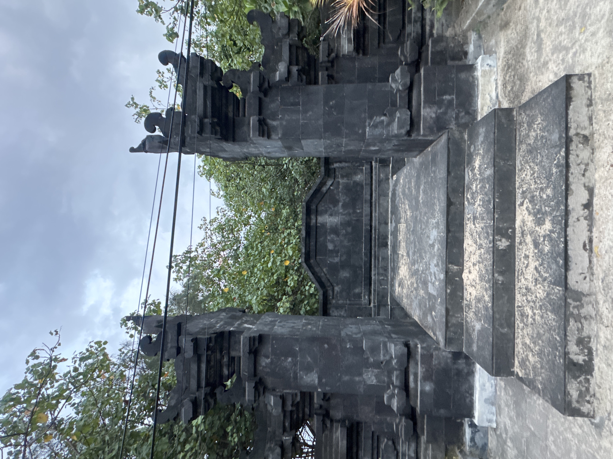 This is a vertical, full-shot, eye-level photograph of a stone gateway in Kuta, Indonesia. The gateway is constructed from dark, rough-hewn stone blocks and features a symmetrical design with two imposing pillars flanking a central staircase. The pillars are elaborately decorated with tiered, carved elements and topped with decorative finials resembling stylized figures. A low stone wall connects the pillars in the background, partially obscured by lush green foliage. Above the gateway, power lines crisscross against a cloudy, overcast sky. The stone steps leading up to the gateway are worn and dusty, indicating foot traffic. The surrounding environment is characterized by dense vegetation, with trees and branches visible on either side of the gateway and peeking over the stone wall. The overall impression is one of age and a traditional architectural style, possibly belonging to a temple or important public space.