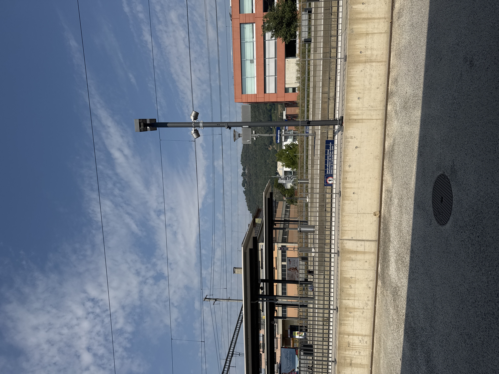 This is a photograph of a train station platform in Mendrisio, Switzerland. The image is taken from ground level, looking towards the train tracks and a modern building. The sky is a bright blue with scattered white clouds.
In the foreground, there is a textured asphalt surface with a circular manhole cover. A dark shadow stretches diagonally across the asphalt from the left side of the frame.
The middle ground features a concrete platform wall topped with a metal fence. Behind the fence, train tracks are visible. To the right of the tracks is a modern, orange building with large, rectangular windows. To the left of the tracks, a covered platform area with a flat roof supported by pillars extends into the scene. Signs are visible on and around the platform infrastructure.
A tall, dark grey pole with two lights and a camera is positioned on the platform near the fence. A sign on this pole reads "Mendrisio."
In the background, a green, tree-covered hillside rises. The overall scene depicts a sunny day at a functional, contemporary train station.