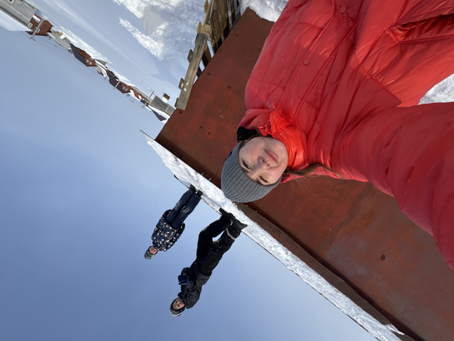 The image captures three individuals in a snow-covered outdoor environment in Gornyi (Dvurechenskoe S/P), Russia, under a clear blue sky. In the immediate foreground, a person wearing a red puffer jacket and a grey knit beanie holds a camera, taking a selfie. Her face is visible and directed towards the camera. Behind her, two other individuals stand on the snow-covered upper surface of a large, rusted, reddish-brown metal structure. The person on the left wears a dark winter jacket, dark pants, and a grey beanie; the person on the right wears a patterned winter jacket, blue jeans, and a grey beanie. Both are looking towards the camera. To the right of the metal structure, partially visible wooden debris is covered in snow. In the distant background, a row of residential houses with red roofs is visible against the snow-covered terrain. The scene is illuminated by natural daylight.